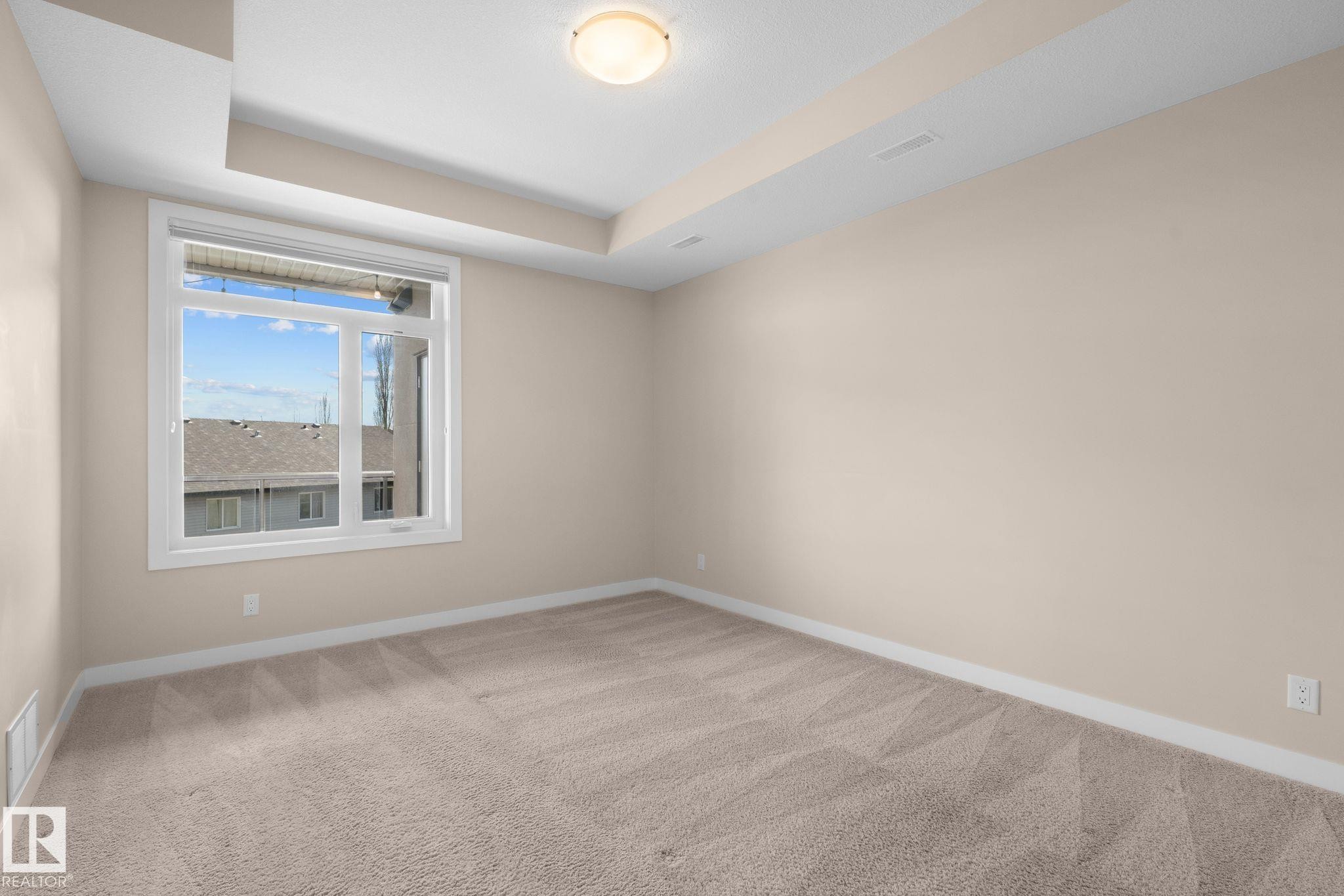 Neutral-toned room featuring a large window, light beige carpeting, tray ceiling, and a flush-mount ceiling light - 309 625 Leger Way, Edmonton, AB