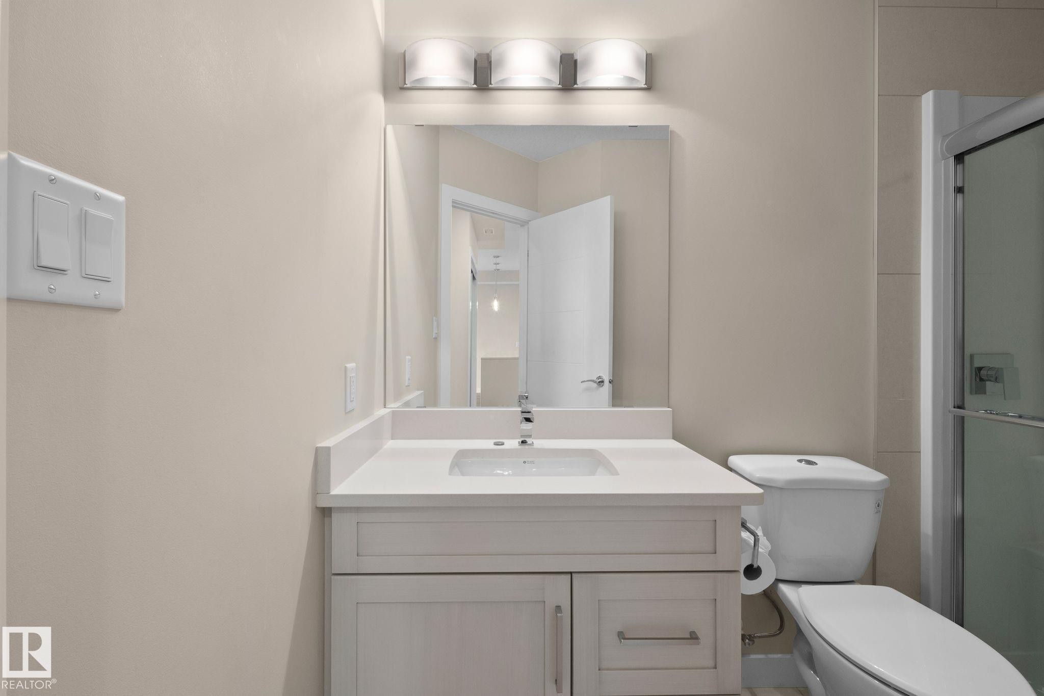 Bathroom vanity with a white countertop and integrated sink, chrome faucet, and a large mirror with a three-light vanity fixture - 309 625 Leger Way, Edmonton, AB