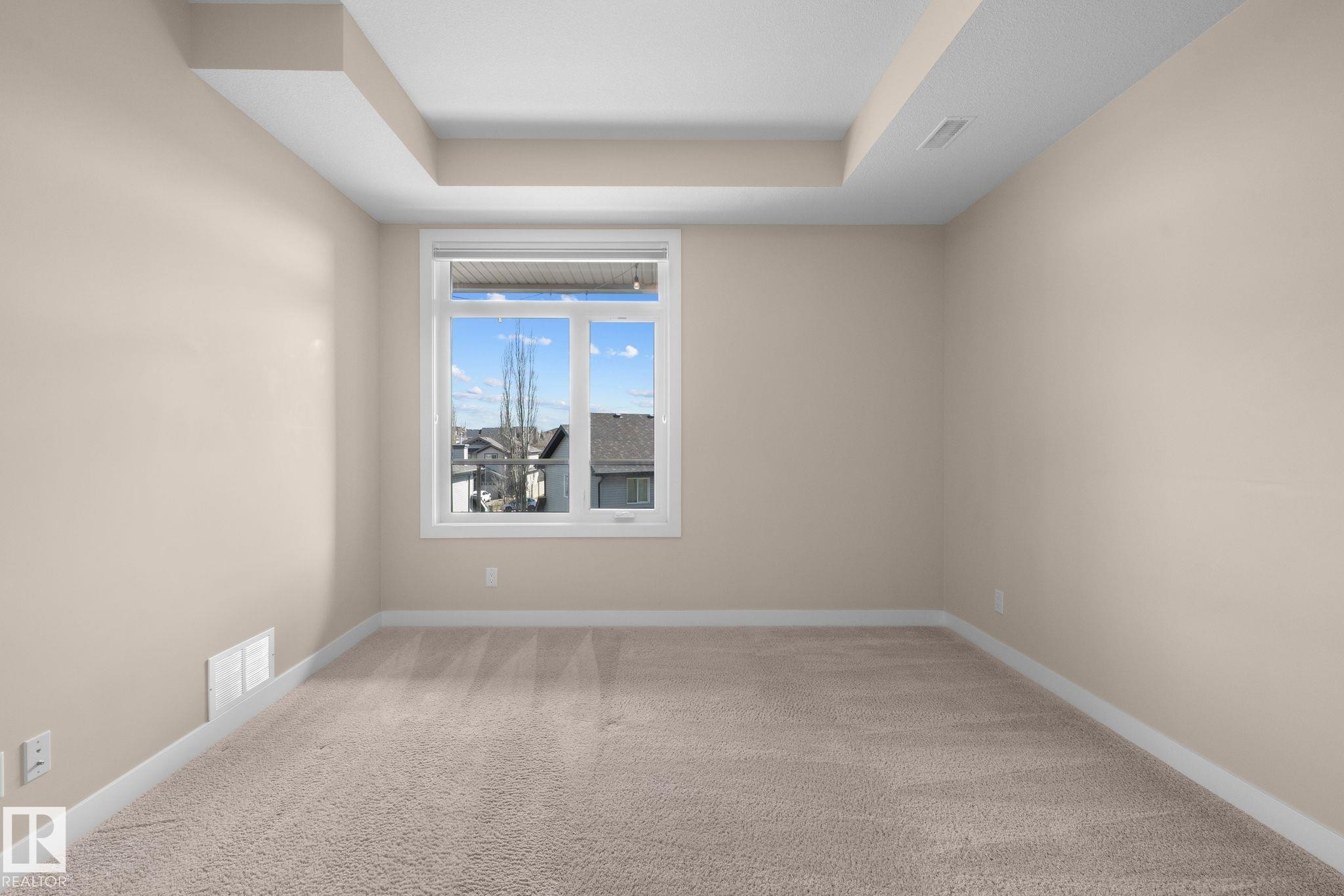 Bright room featuring a large window, neutral wall paint, tray ceiling, plush carpeting, and white baseboards - 309 625 Leger Way, Edmonton, AB