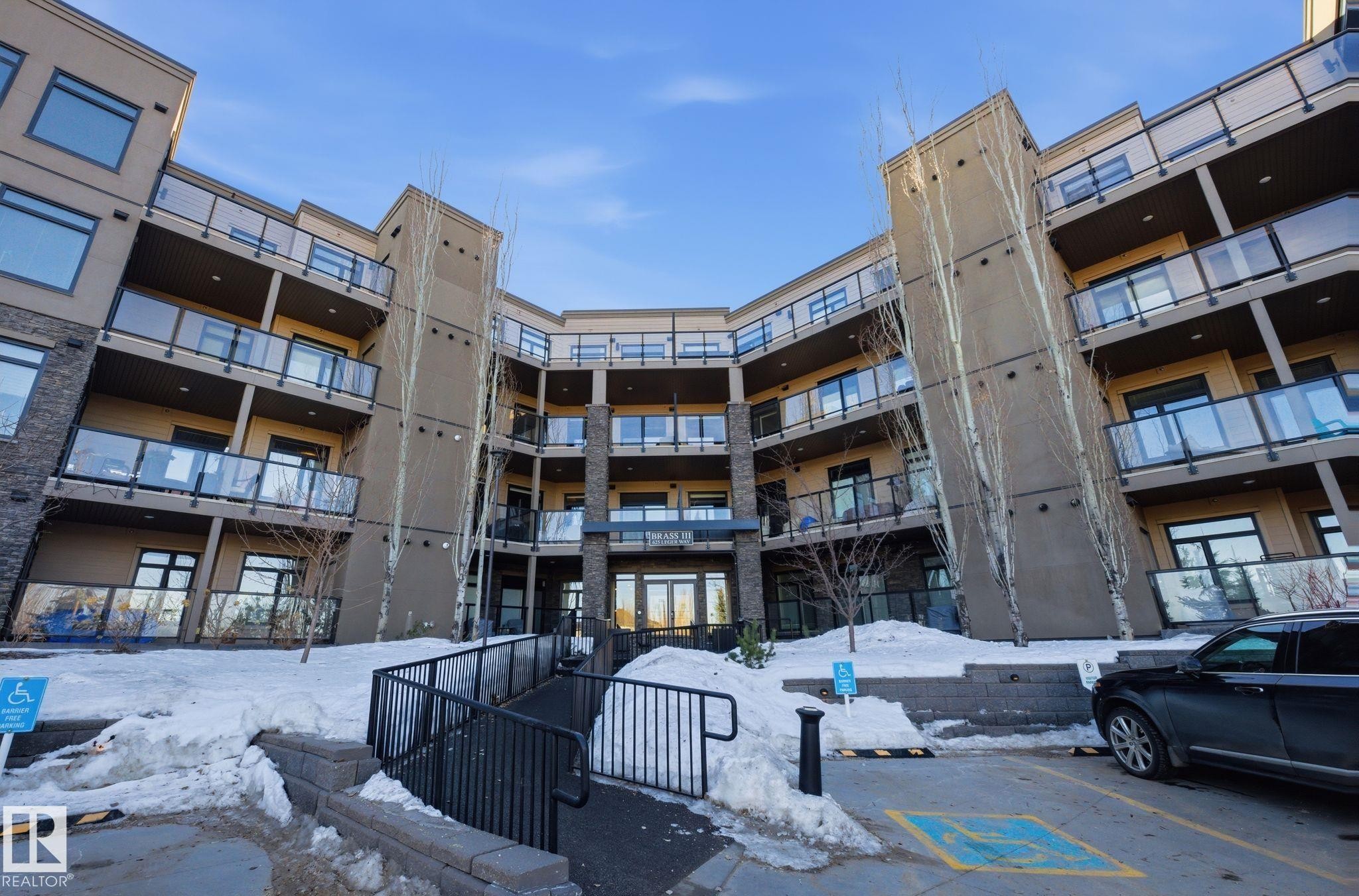 Contemporary multi-story building featuring warm-toned siding, dark brick accents, glass-paneled balconies, and a central entry with a ramp - 309 625 Leger Way, Edmonton, AB