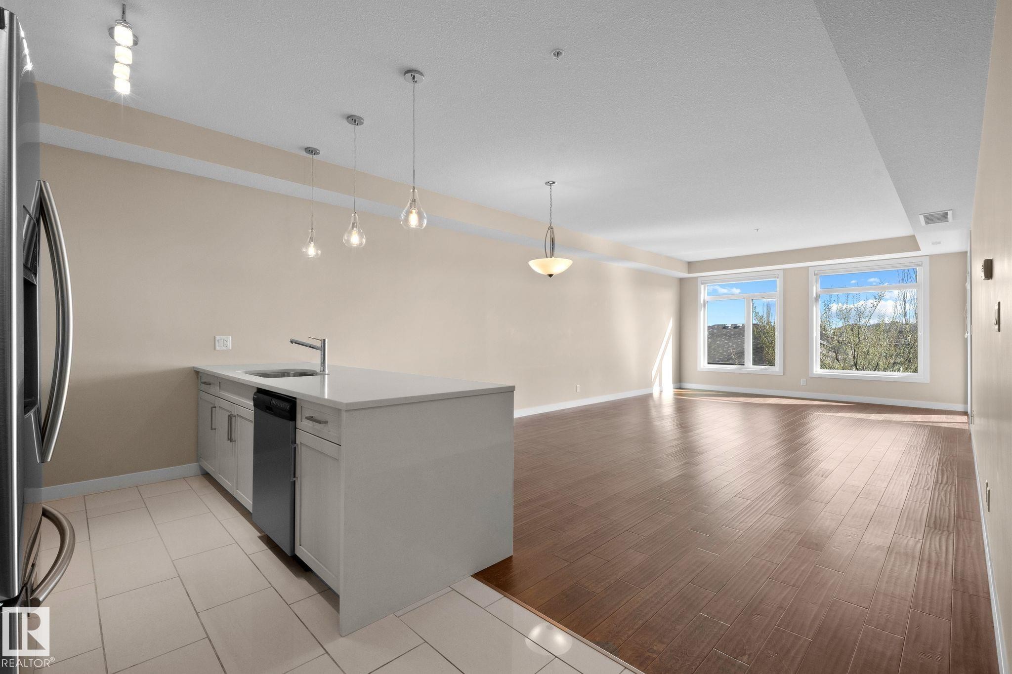Open-concept living area featuring wood-finish flooring, a kitchen island with a built-in sink and dishwasher, pendant lighting, and multiple windows - 309 625 Leger Way, Edmonton, AB