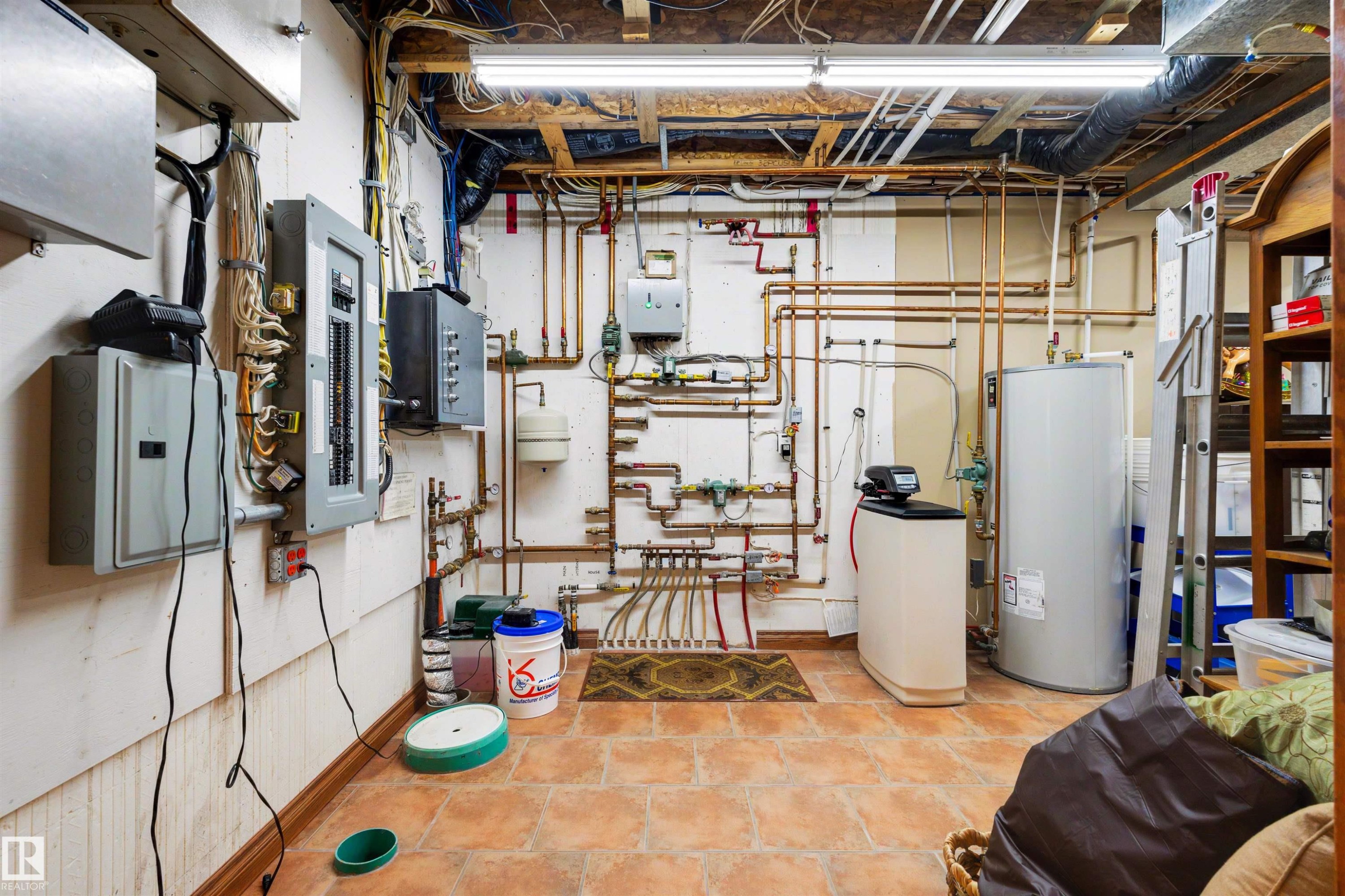 Utility room featuring exposed ceiling joists, overhead strip lighting, multiple electrical panels, extensive copper piping, and a water heater - 28125 Highway 587, Rural Red Deer County, AB - Indoor Photo Showing Basement