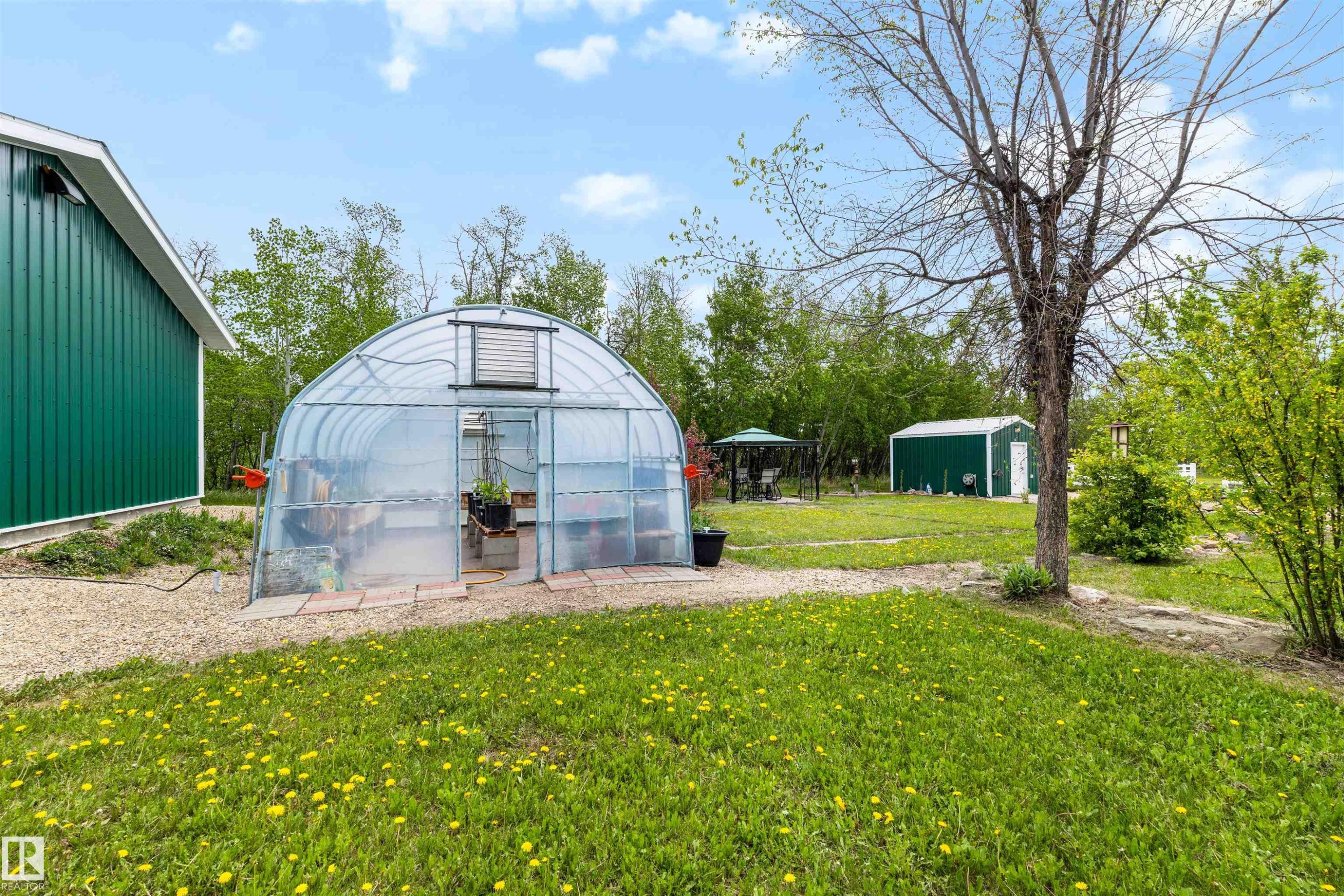 Expansive outdoor area featuring a prominent greenhouse with a clear arched roof, a large green outbuilding with vertical siding, and a smaller green shed - 28125 Highway 587, Rural Red Deer County, AB - Outdoor