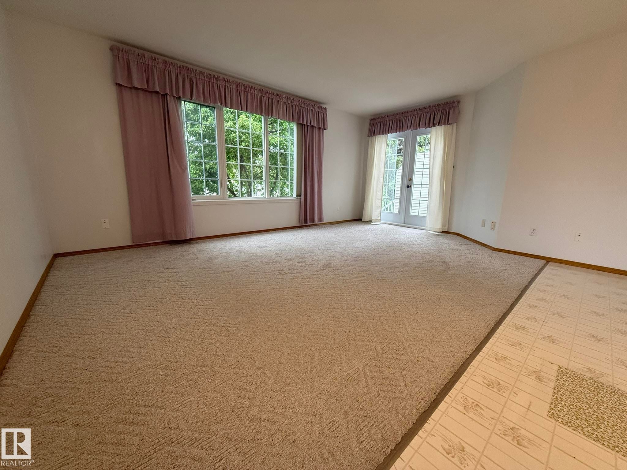 Spacious room featuring neutral carpeting, white walls, a large multi-pane window, and white French doors - 102 4610 50 Avenue, Stony Plain, AB