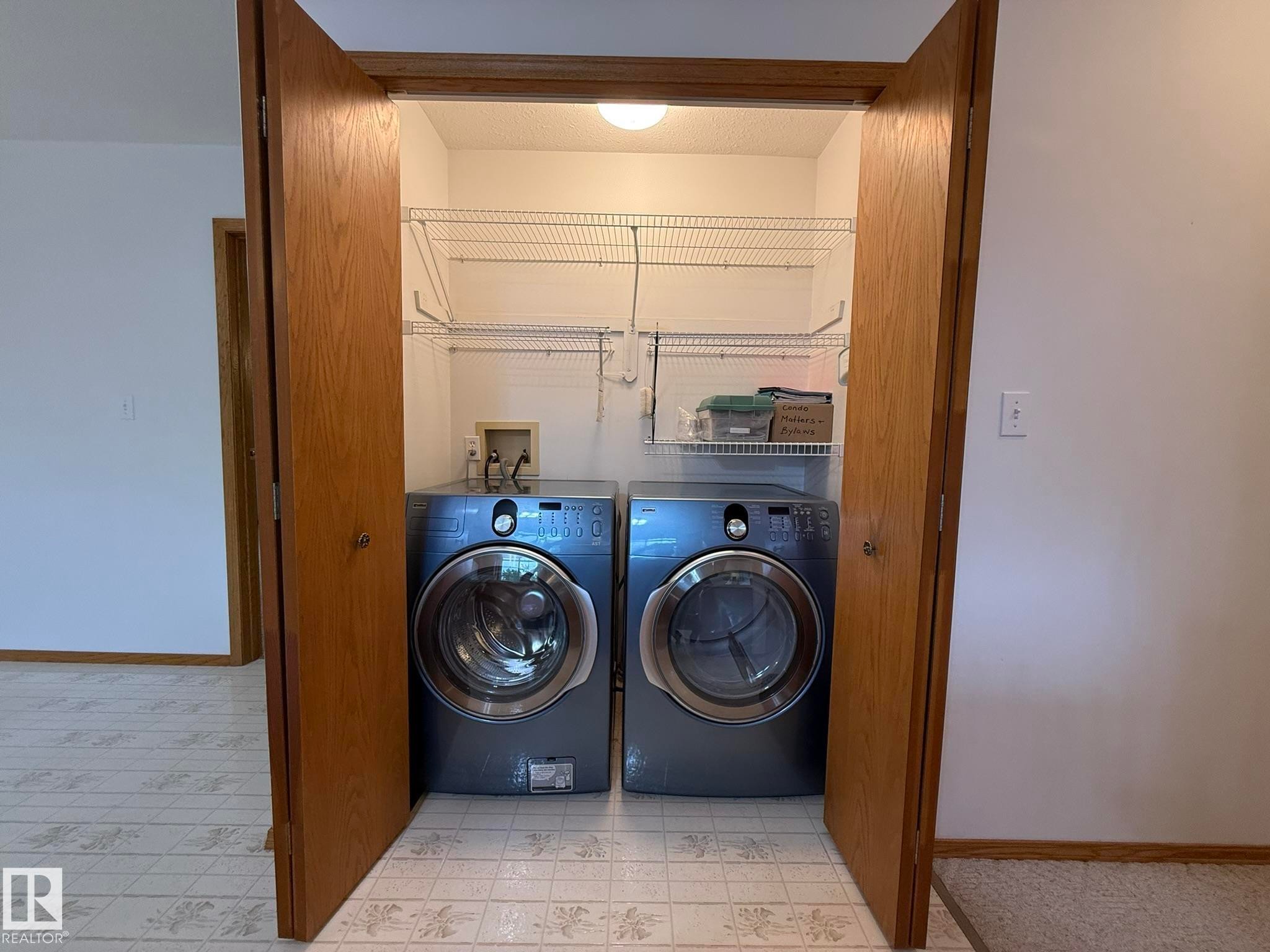 Dedicated laundry area featuring bi-fold doors, upper wire shelving, and a patterned floor - 102 4610 50 Avenue, Stony Plain, AB