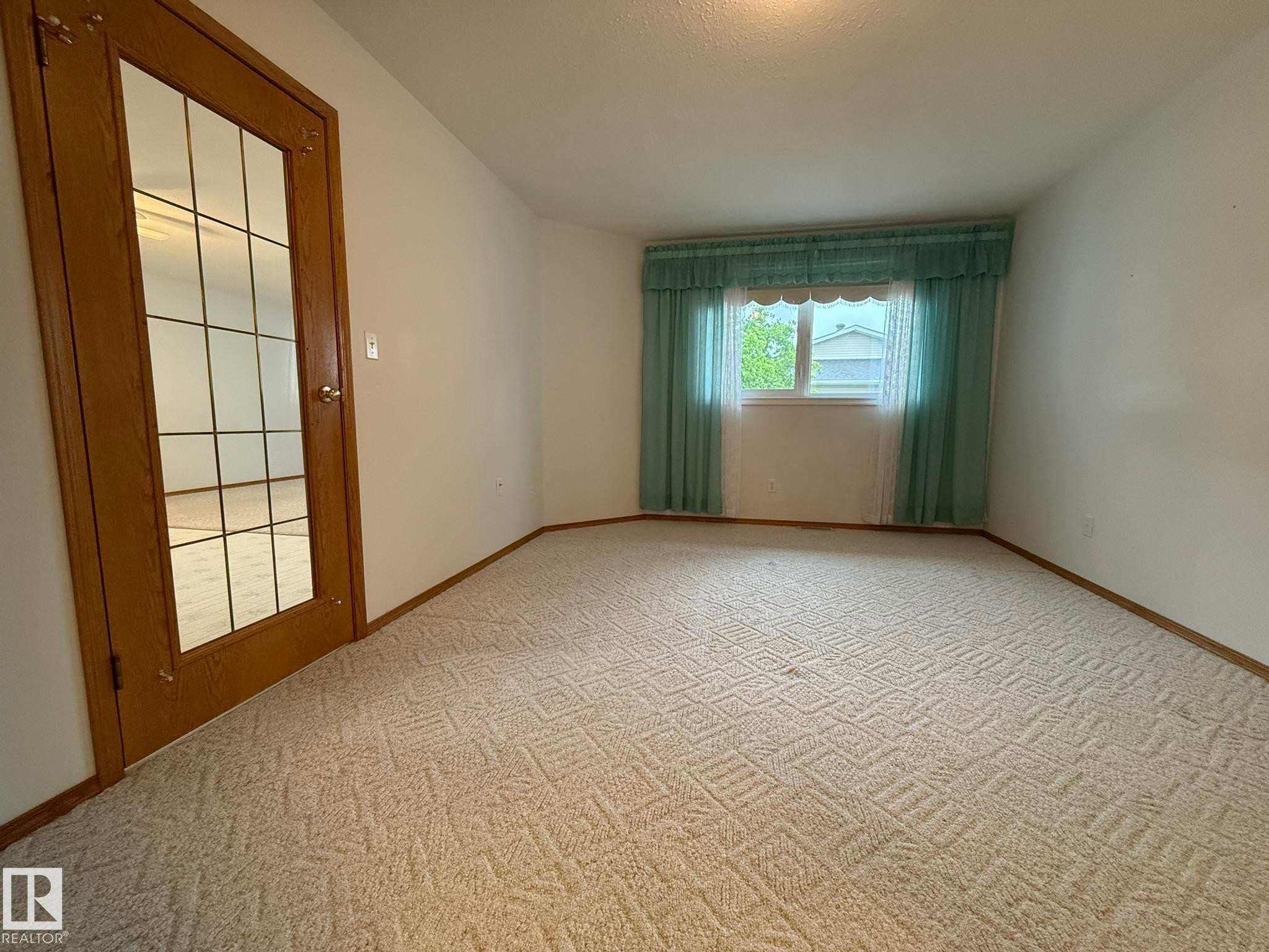 Window with light blue drapes and valance, light-toned patterned carpeting, white walls, and a wood-finish door with glass panels - 102 4610 50 Avenue, Stony Plain, AB