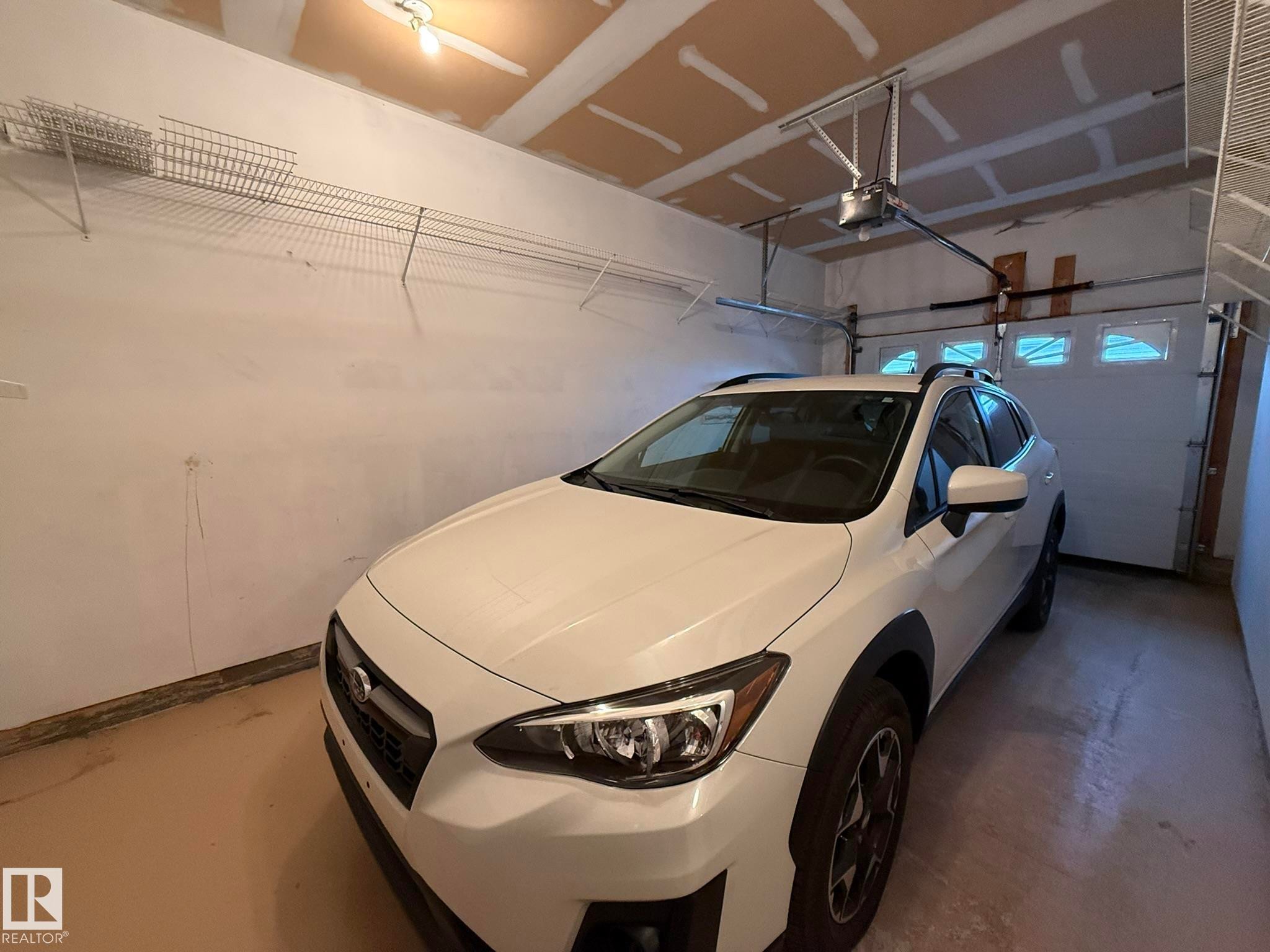 Attached garage featuring a light brown floor finish, white walls, and exposed ceiling joists - 102 4610 50 Avenue, Stony Plain, AB