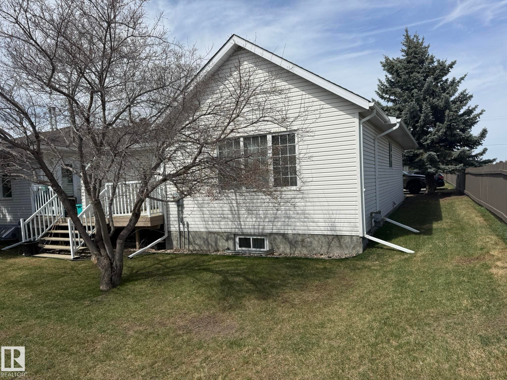Residential exterior featuring light-colored horizontal siding, a gabled roof, and a mature evergreen tree - 102 4610 50 Avenue, Stony Plain, AB