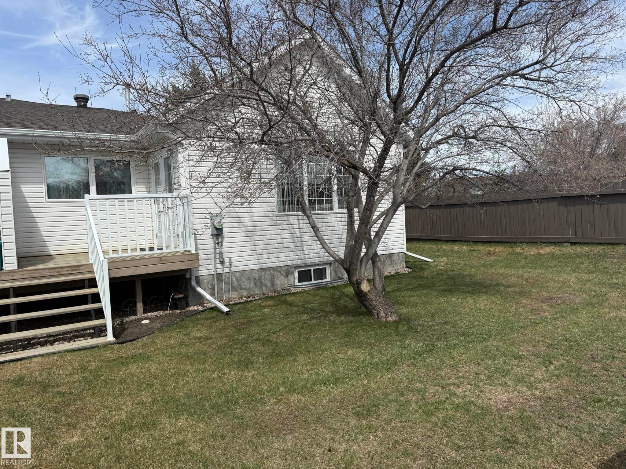 Rear exterior featuring white vinyl siding, a wood deck with white railings, a mature deciduous tree, and a brown privacy fence - 102 4610 50 Avenue, Stony Plain, AB