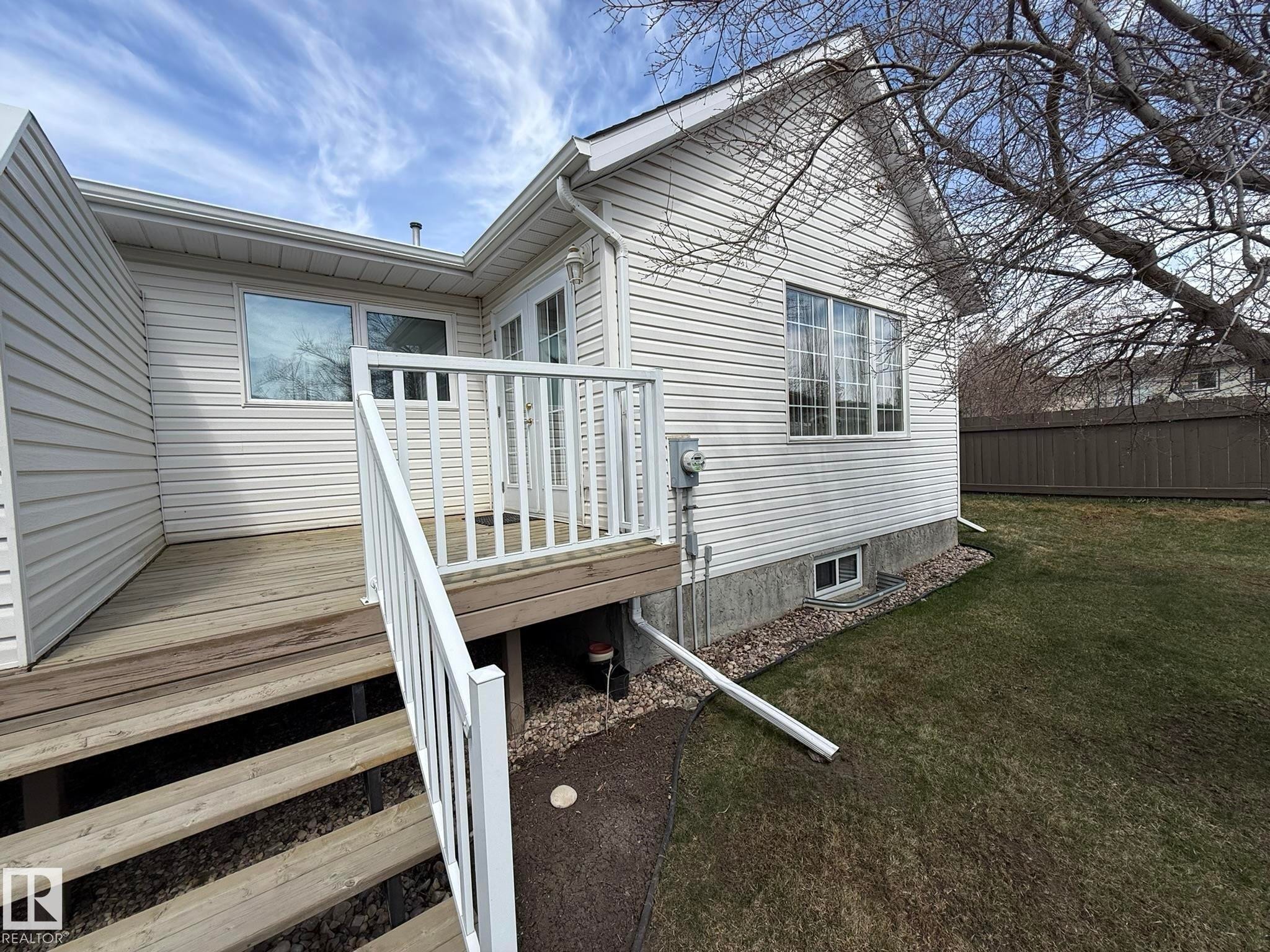 Rear exterior featuring a wooden deck with white railing, vinyl siding, multiple windows, and a grassy yard with a privacy fence - 102 4610 50 Avenue, Stony Plain, AB