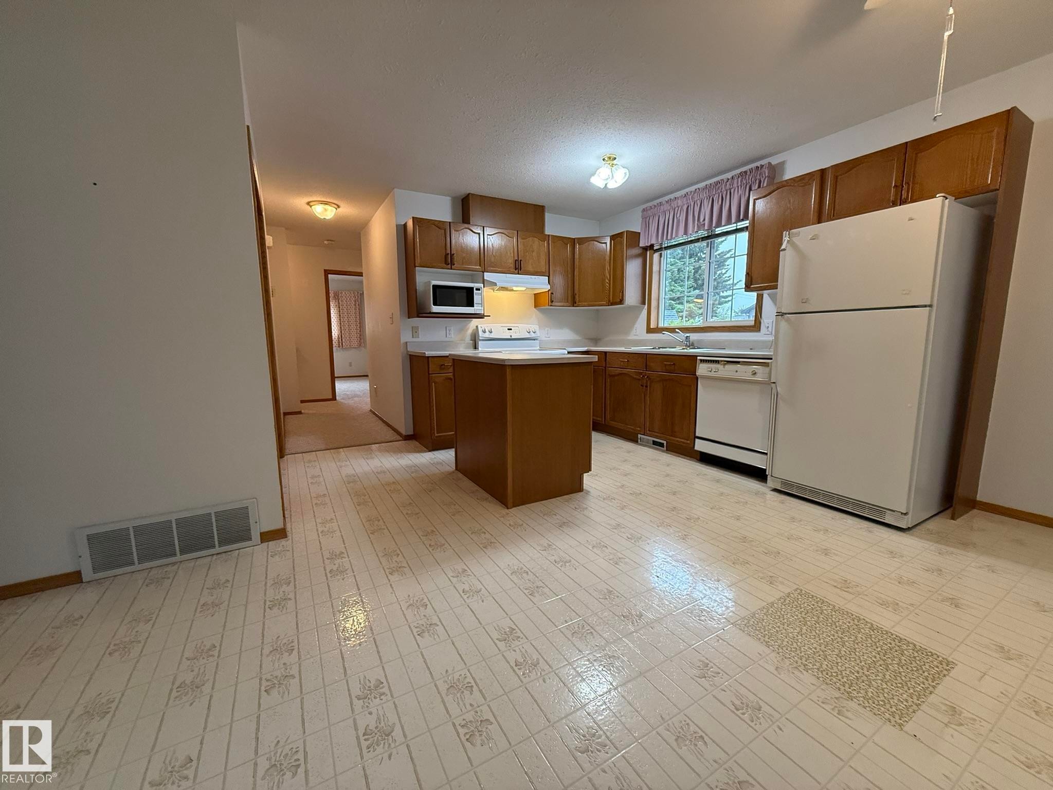 Kitchen featuring wood cabinetry, white appliances, a center island with countertop, and patterned flooring - 102 4610 50 Avenue, Stony Plain, AB