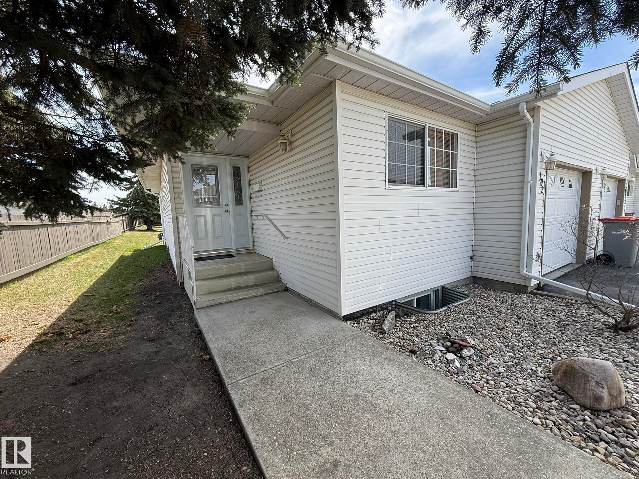 White vinyl siding exterior with an attached garage, concrete walkway, and entryway steps - 102 4610 50 Avenue, Stony Plain, AB