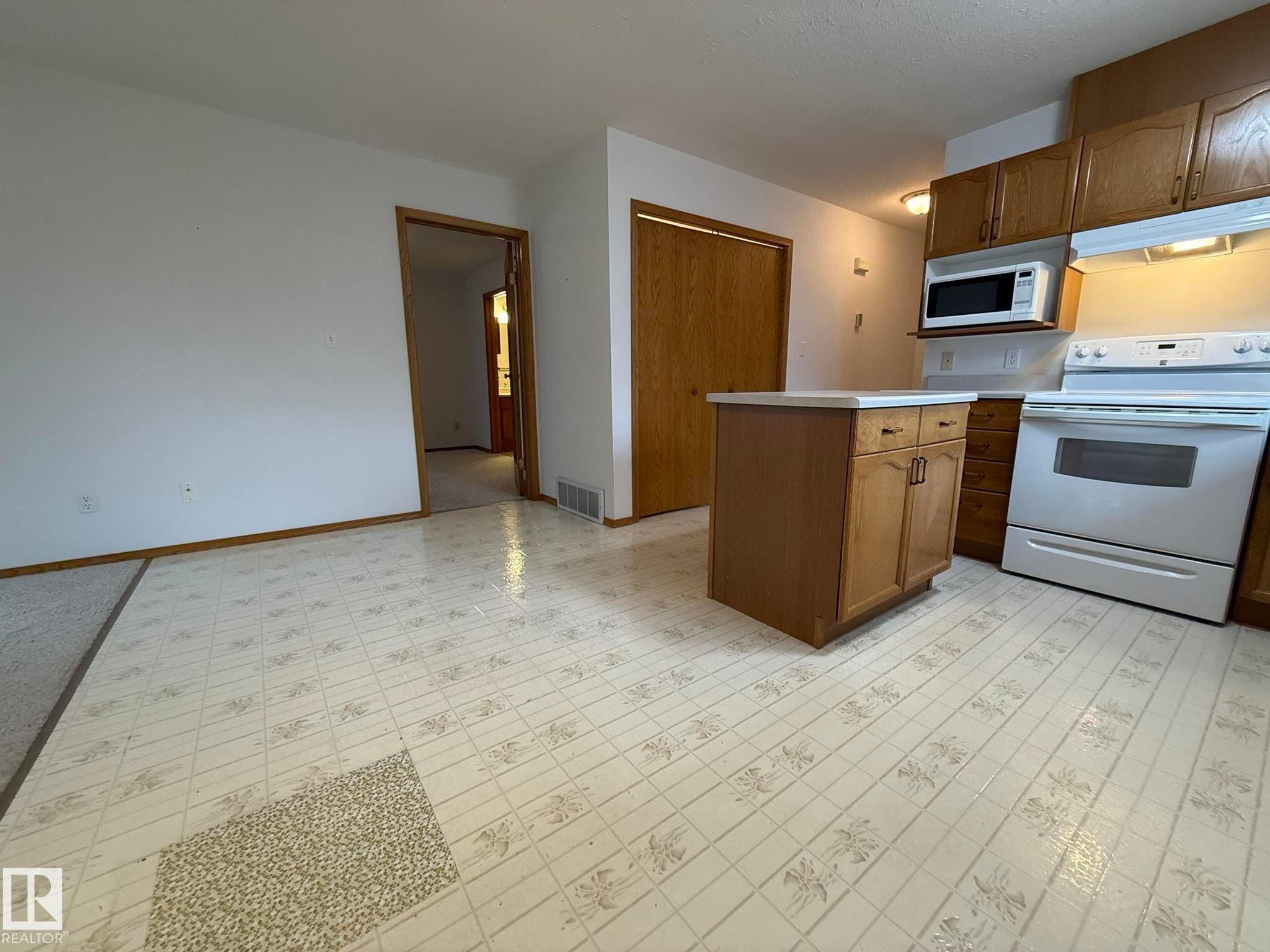 Kitchen featuring a freestanding white range, white microwave, wood-finish cabinetry, and a central island with storage and a light-colored countertop - 102 4610 50 Avenue, Stony Plain, AB
