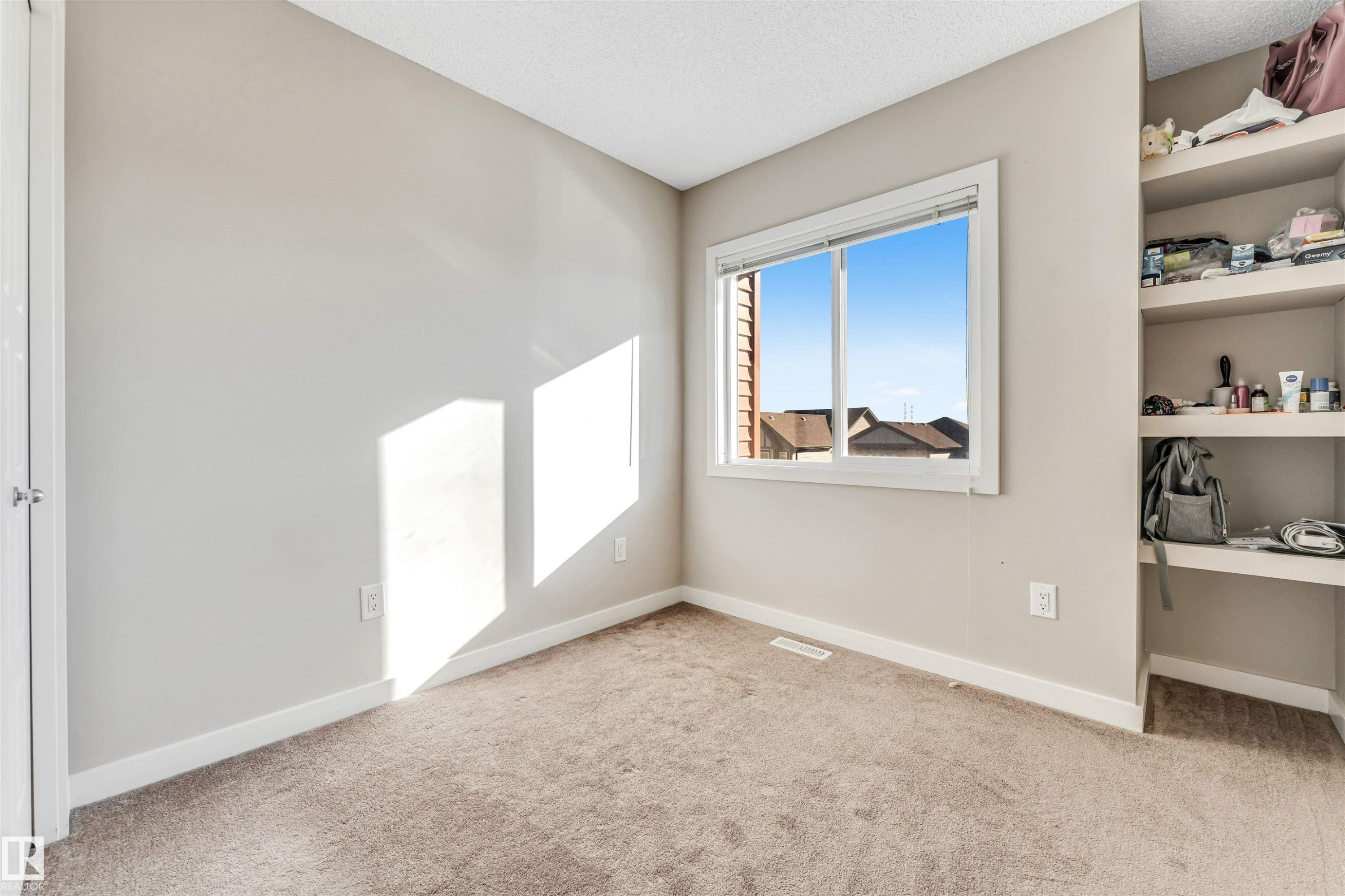 Neutral-toned room featuring a window with white trim, built-in shelving, light brown carpeting, and white baseboards - 1624 33A Street, Edmonton, AB