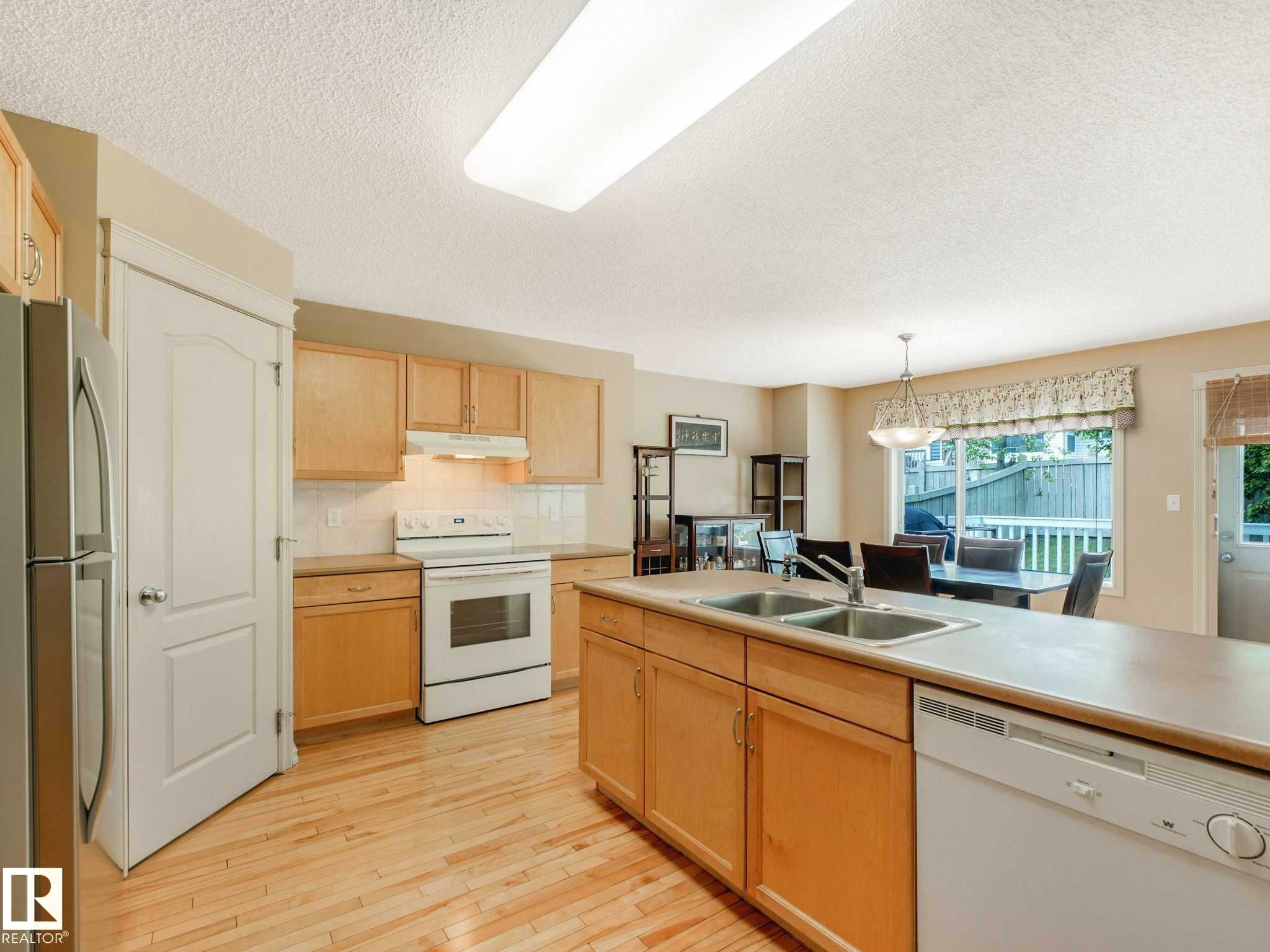 Kitchen featuring wood-finish flooring, light wood cabinetry, white appliances, and an island with a double basin sink - 1116 Lincoln Crescent, Edmonton, AB - Indoor Photo Showing Kitchen With Double Sink