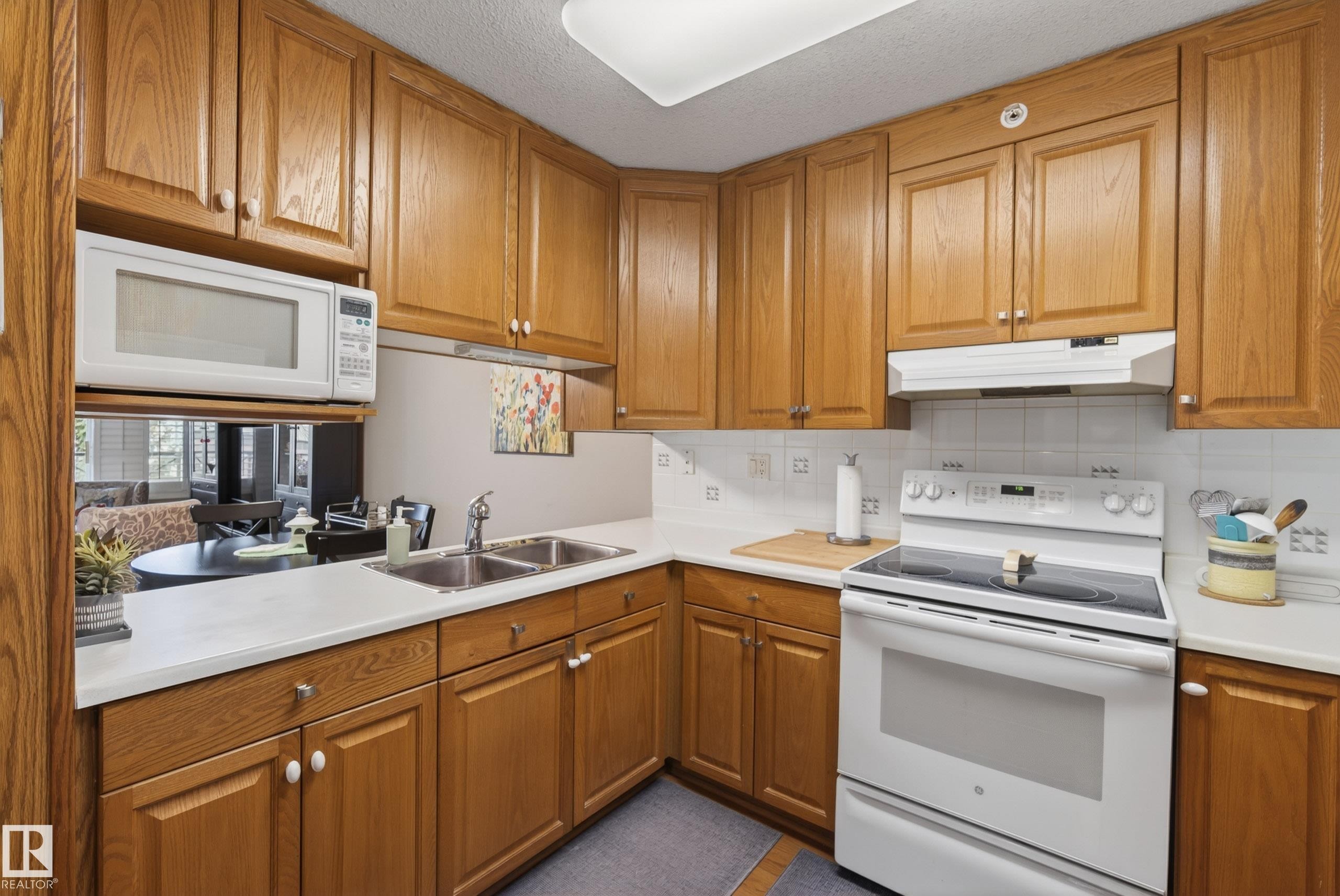 Kitchen featuring wood cabinetry, white countertops, dual basin stainless steel sink, and white tile backsplash - 413 8912 156 Street Nw, Edmonton, AB