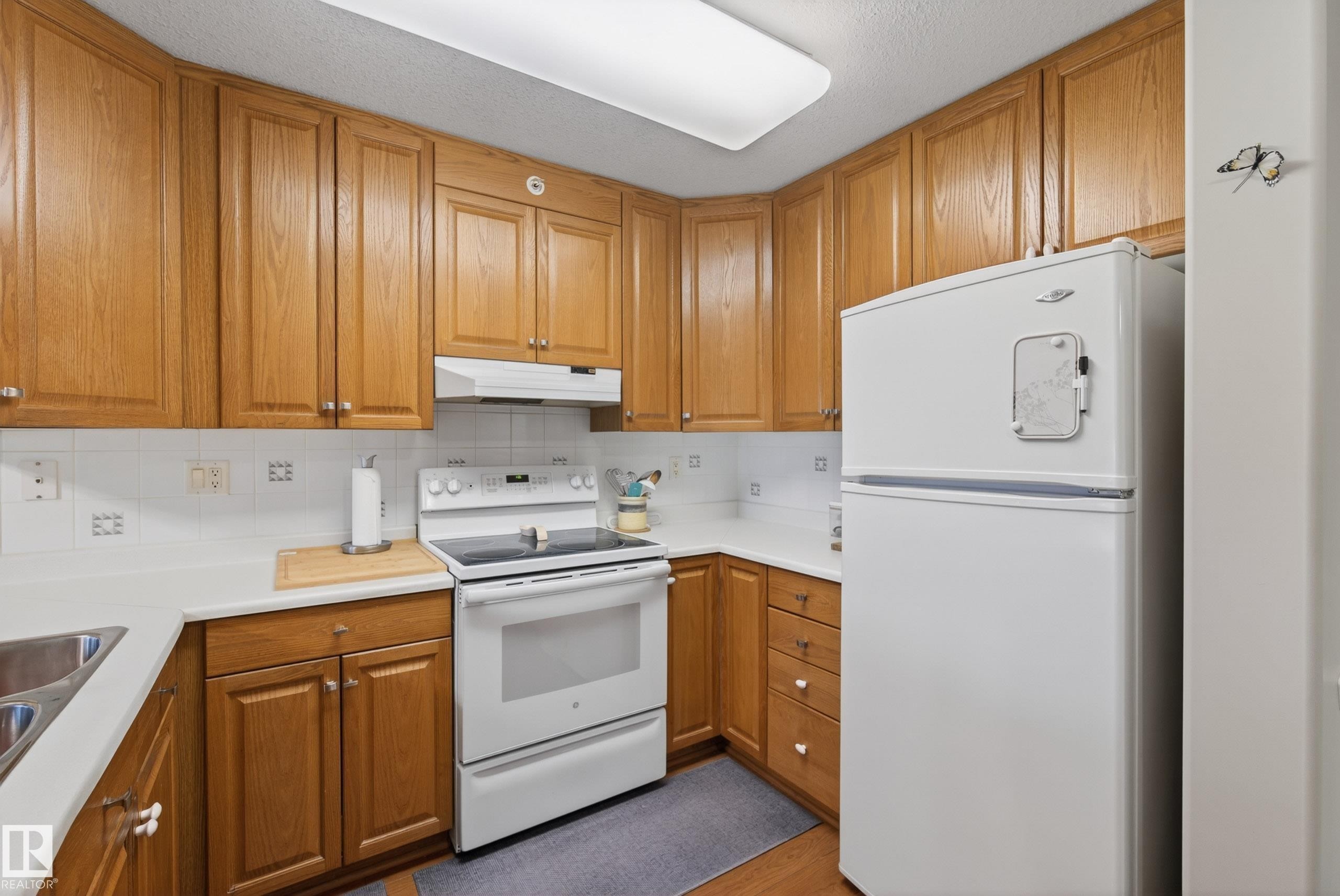 Kitchen featuring wood cabinetry, white countertops, tile backsplash, stainless steel sink, and white appliances - 413 8912 156 Street Nw, Edmonton, AB