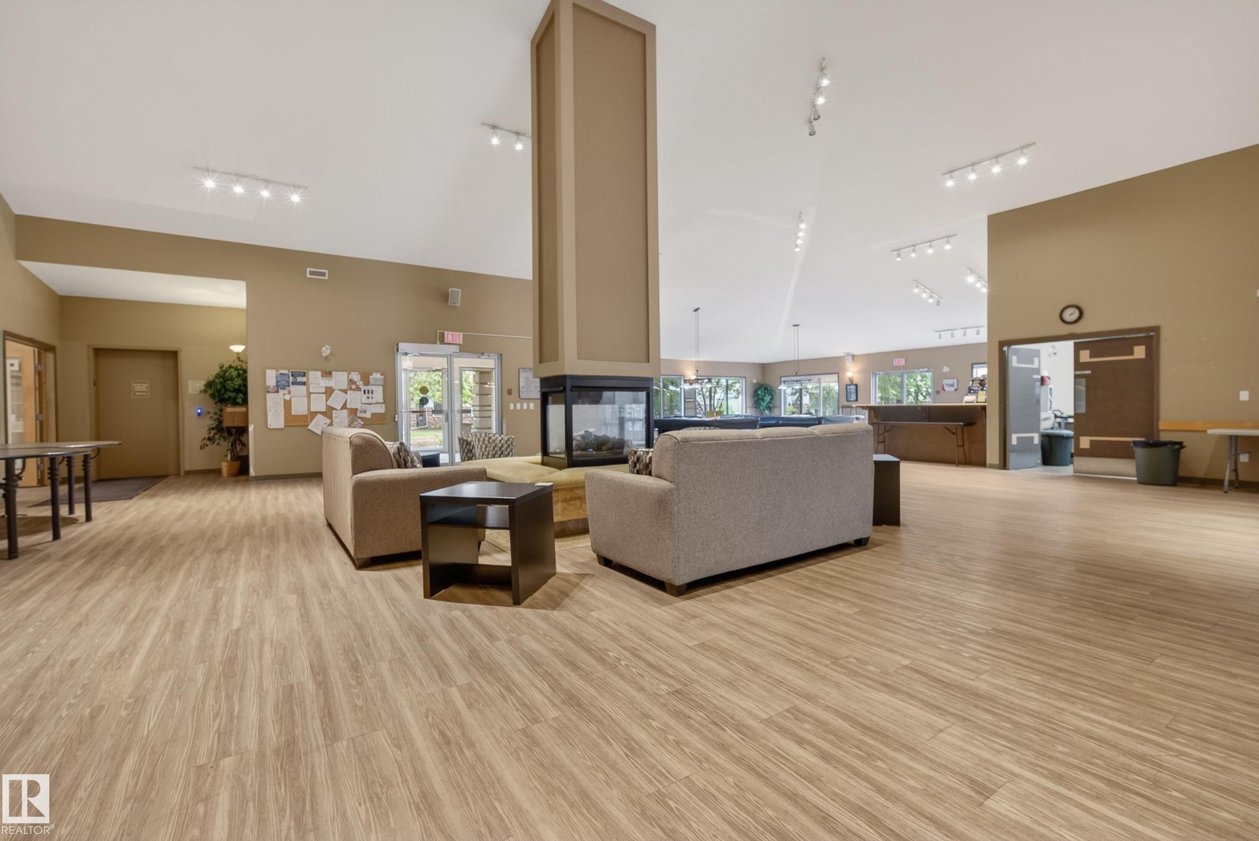 Spacious common area featuring vaulted ceilings, wood-finish flooring, a central three-sided fireplace, recessed lighting, and multiple windows - 413 8912 156 Street Nw, Edmonton, AB