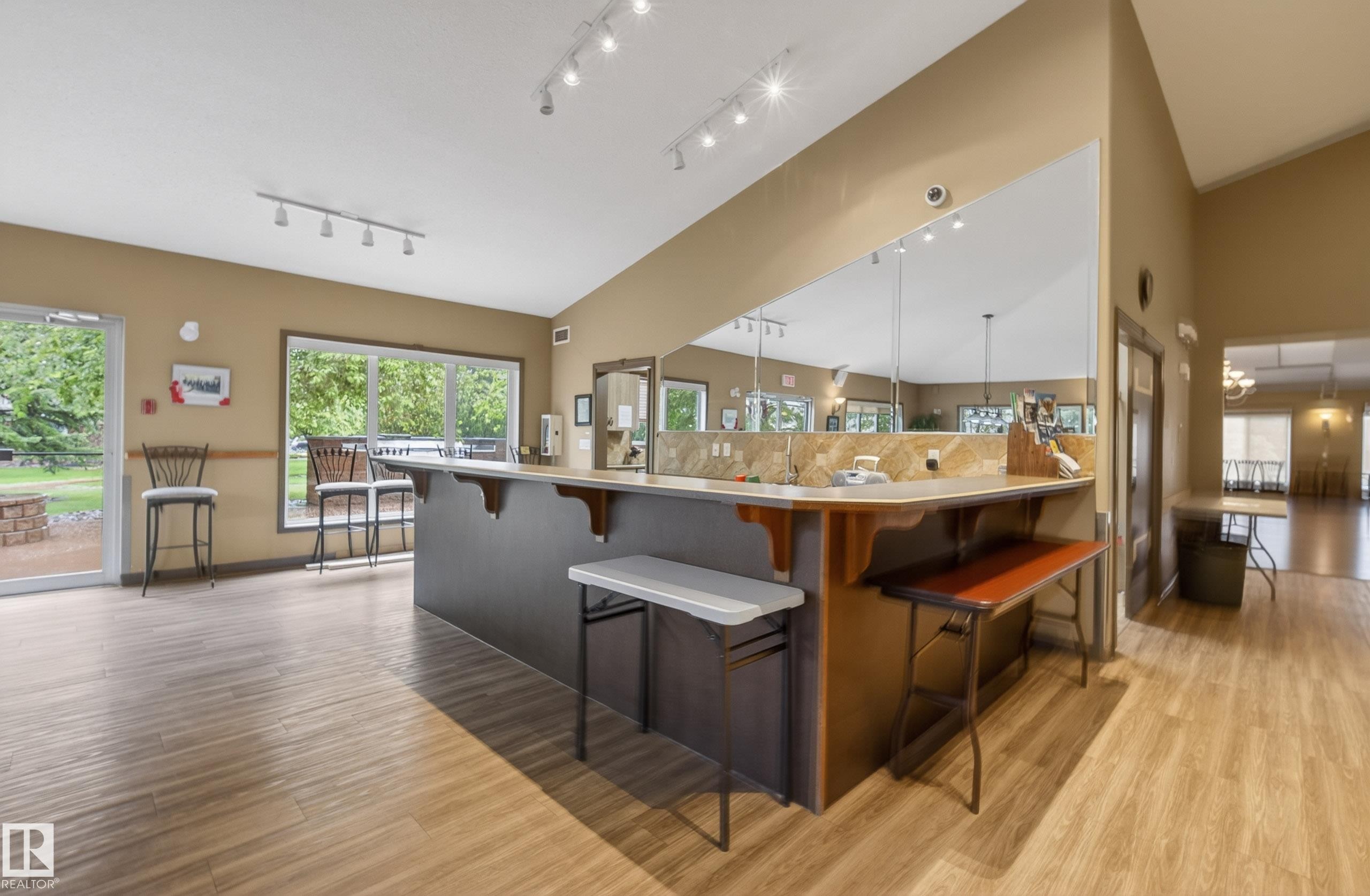 Open-concept common area featuring wood-finish flooring, a large service bar with stone-look countertop and backsplash, track lighting, and expansive windows - 413 8912 156 Street Nw, Edmonton, AB