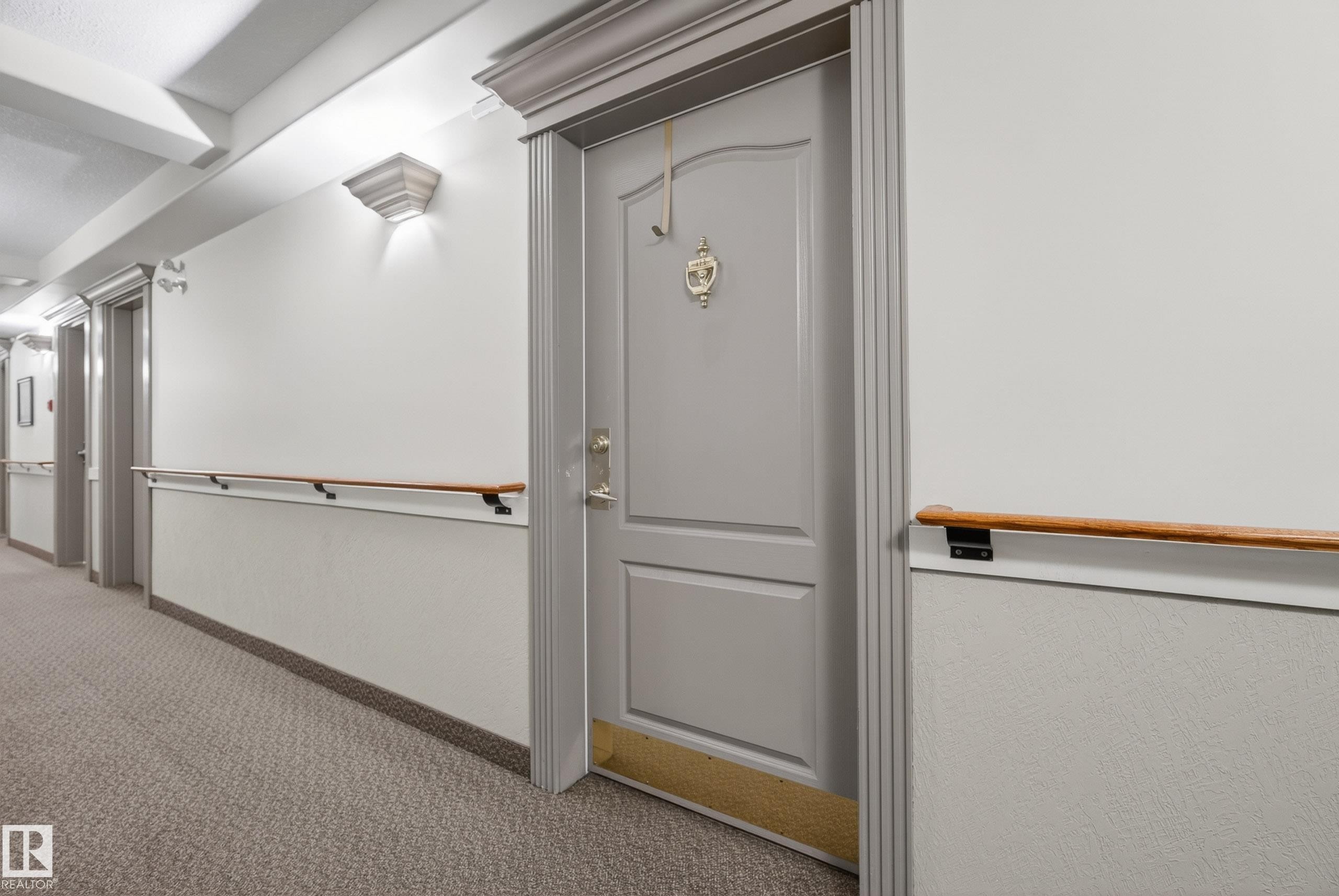 Hallway featuring wall sconces, textured lower wall paneling, and a wood handrail - 413 8912 156 Street Nw, Edmonton, AB