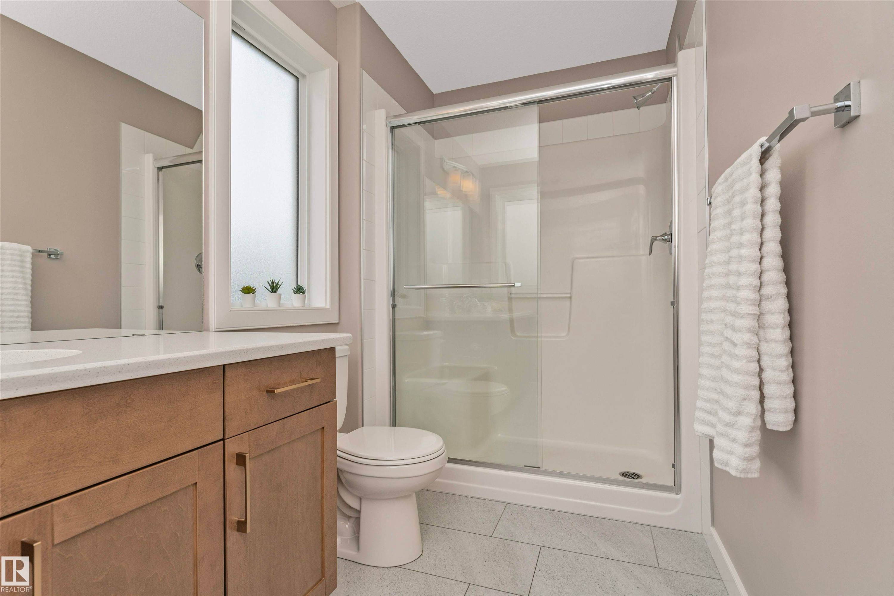 Bathroom featuring a full shower enclosure with sliding glass doors, a single vanity with wood-finish cabinetry, a white countertop with an undermount sink, and light gray tile flooring - 22124 85 Avenue Nw, Edmonton, AB - Indoor Photo Showing Bathroom