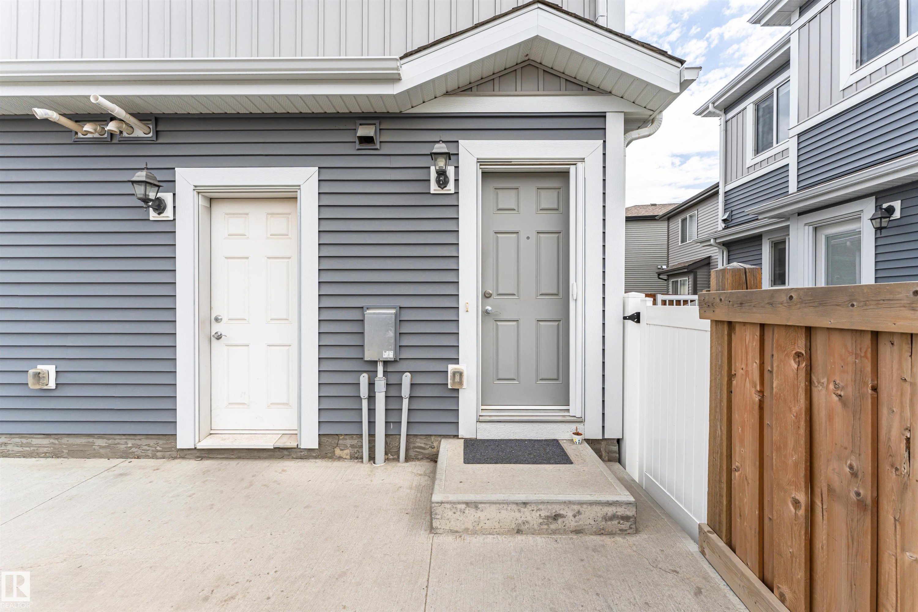Exterior featuring grey horizontal siding, a light grey entry door with white trim, a white utility door with white trim, a concrete patio, and a wood and white privacy fence - 22124 85 Avenue Nw, Edmonton, AB - Outdoor