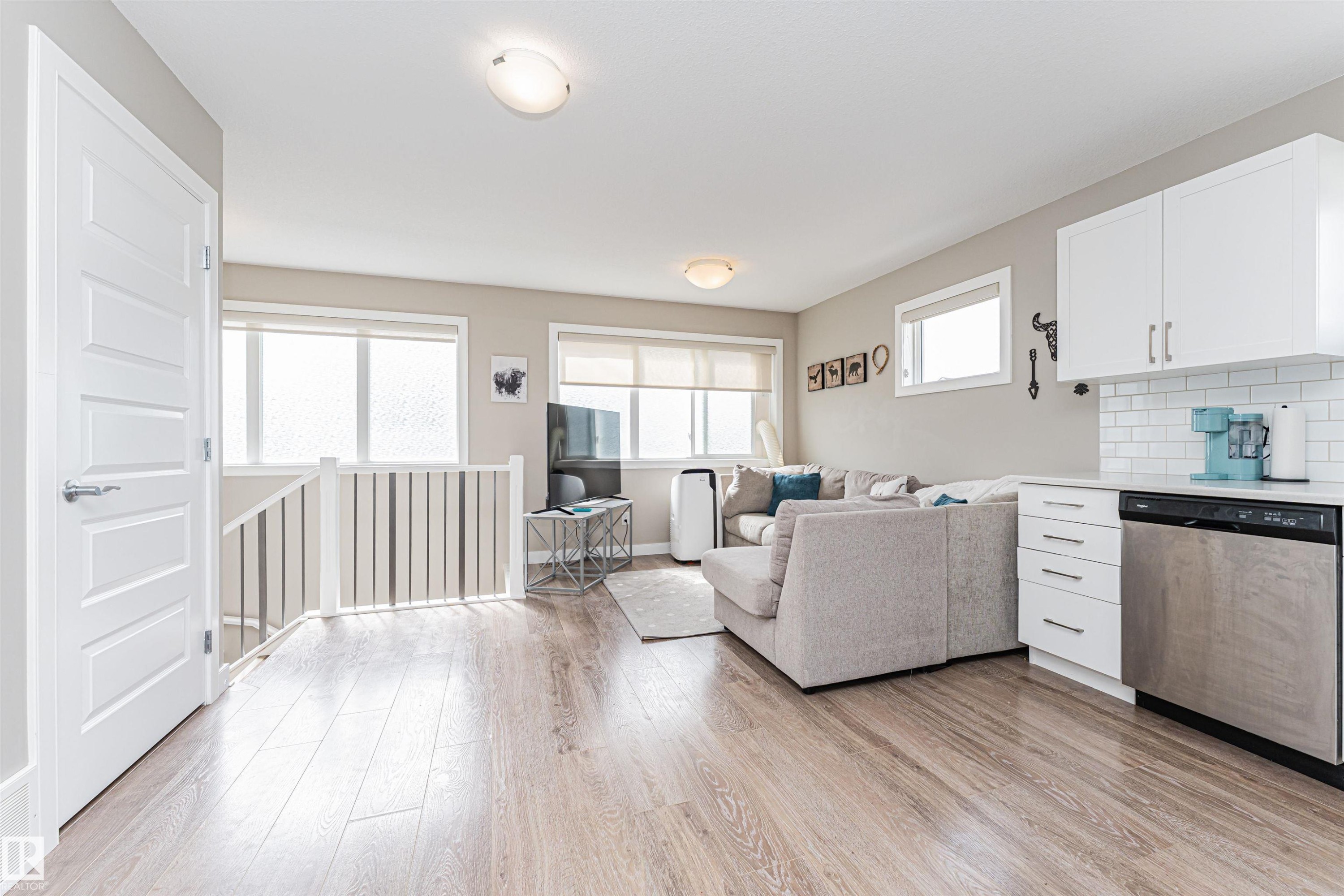 Open-concept living space featuring wood-finish flooring, white shaker cabinetry, stainless steel dishwasher, white subway tile backsplash, and light neutral wall paint - 22124 85 Avenue Nw, Edmonton, AB - Indoor Photo Showing Other Room