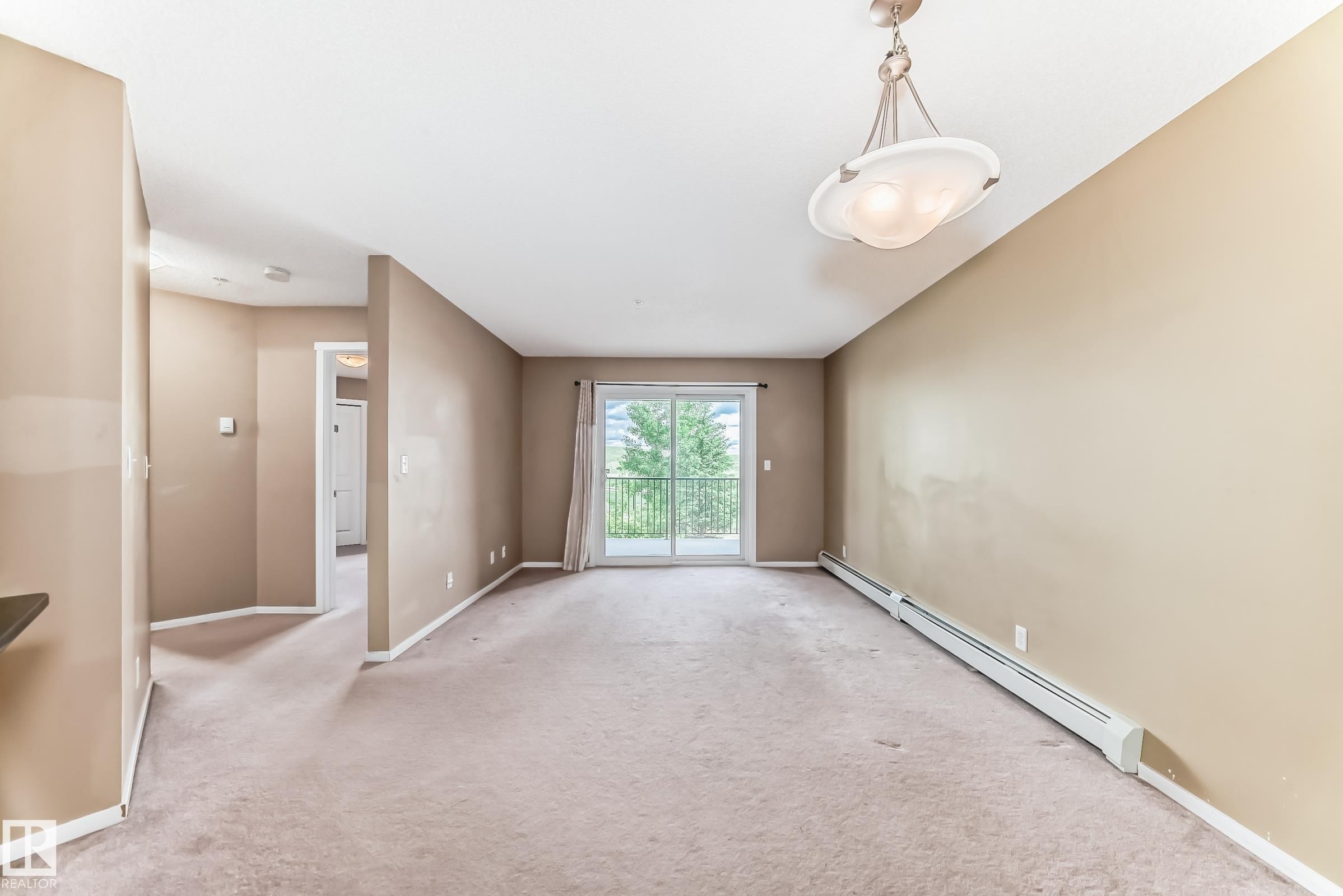 Living area featuring a glass sliding door to a balcony, a ceiling-mounted light fixture, neutral carpeting, and baseboard heating - 215 1510 Watt Drive, Edmonton, AB