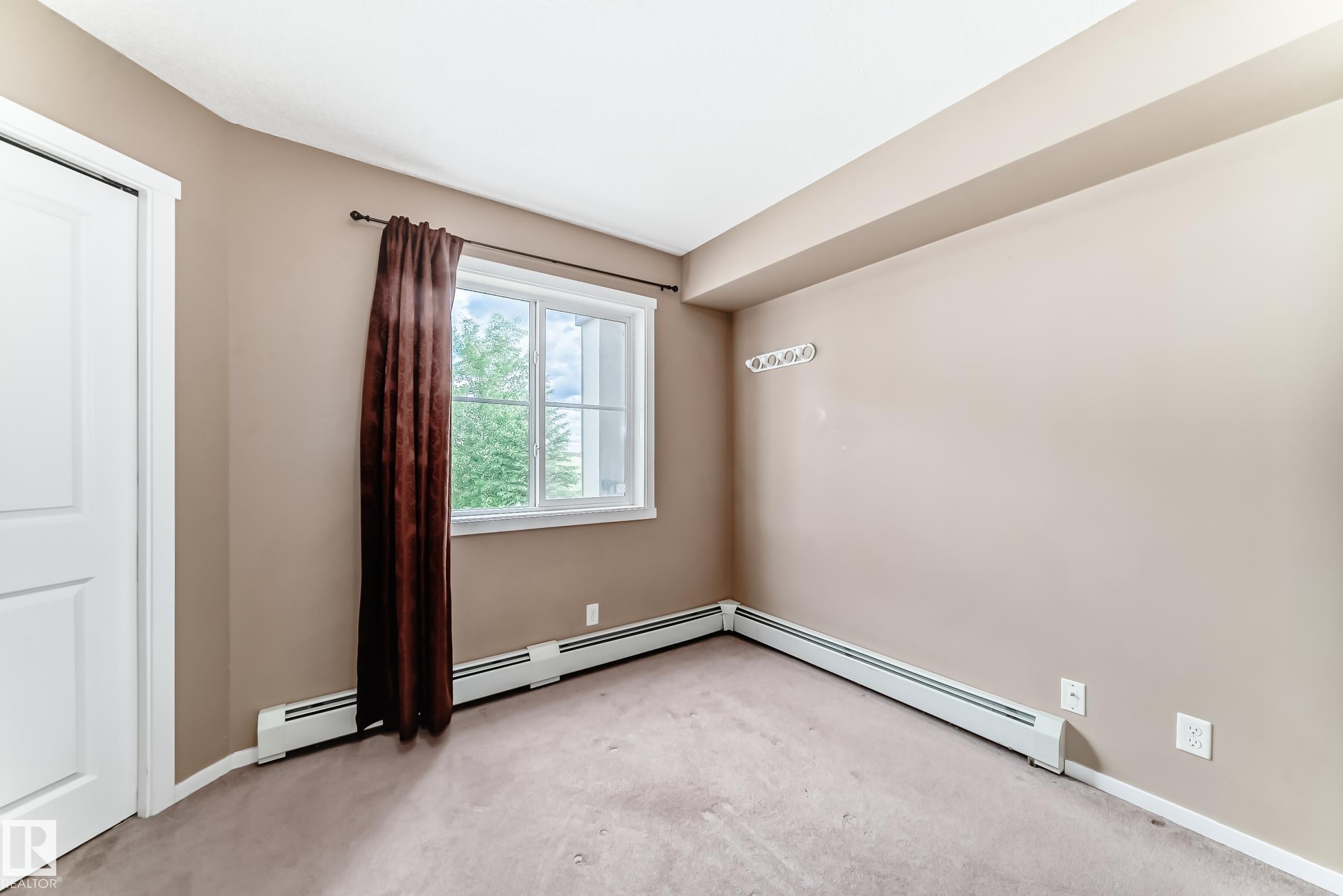 Carpeted interior space featuring a white door, large window with white trim, baseboard heating, and light tan wall color - 215 1510 Watt Drive, Edmonton, AB