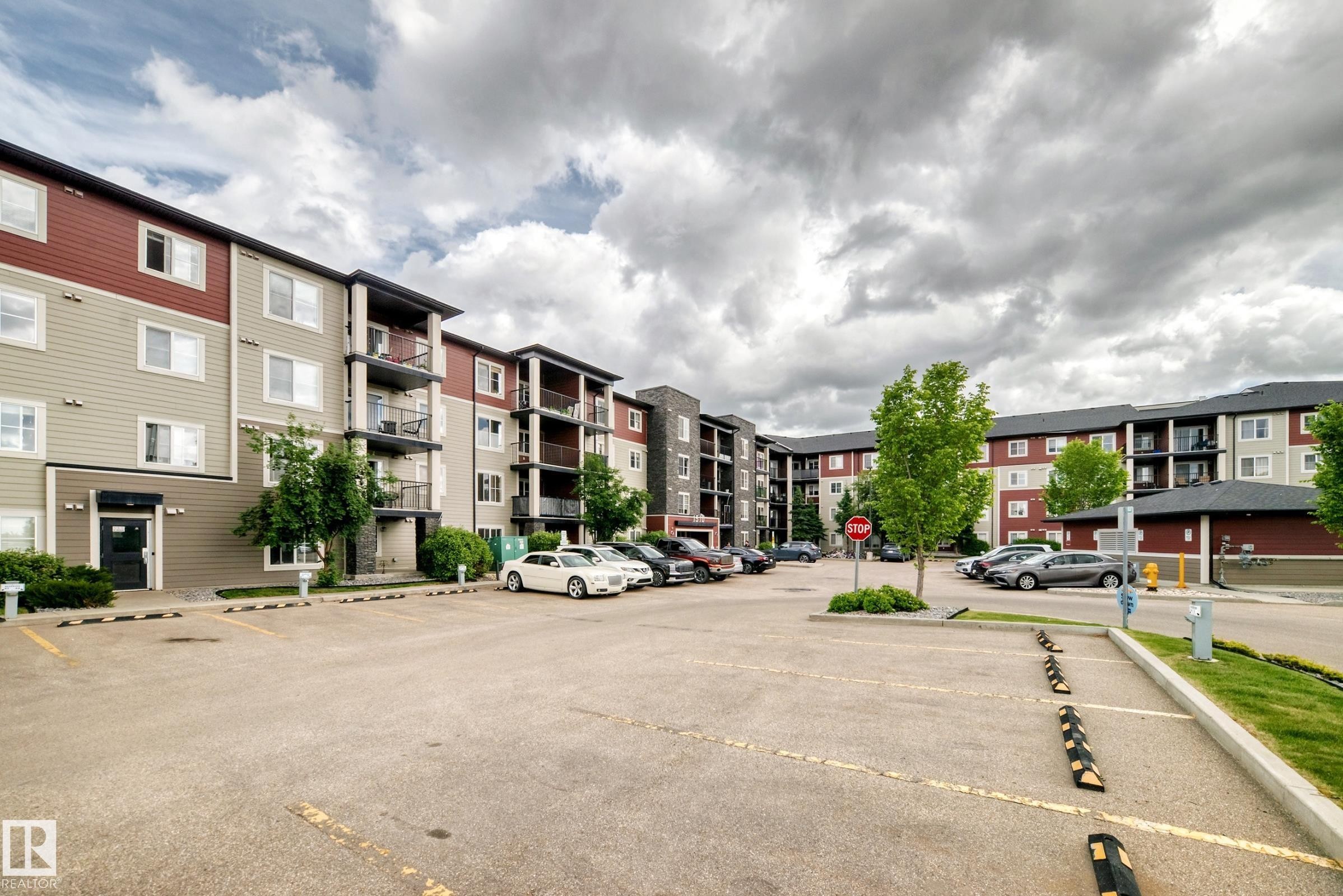 Multi-story residential building featuring horizontal siding in a two-tone color scheme, individual balconies with dark railings, multiple large windows, and a paved parking area - 215 1510 Watt Drive, Edmonton, AB