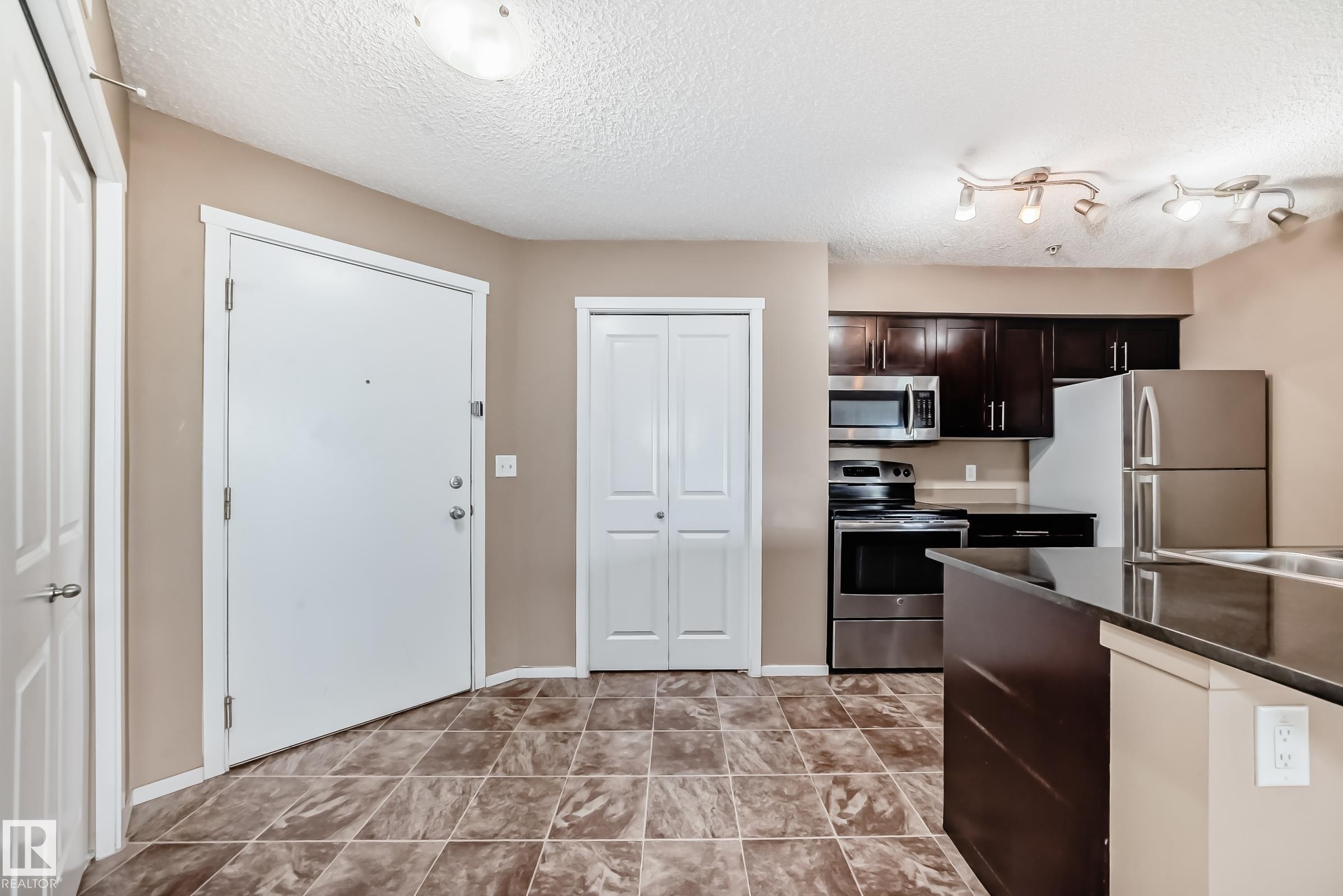 Open-concept kitchen featuring dark cabinetry, stainless steel appliances, a dark countertop, and track lighting - 215 1510 Watt Drive, Edmonton, AB
