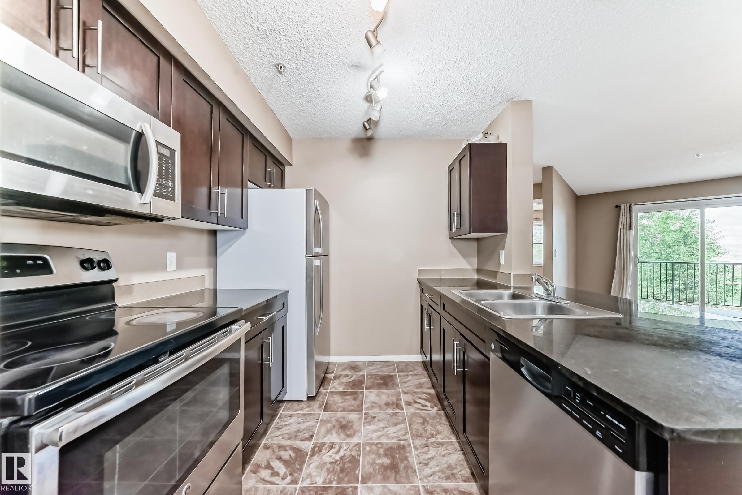 Kitchen featuring dark wood-finish cabinetry, stainless steel appliances, dark countertops, a dual-basin sink, and tiled flooring - 215 1510 Watt Drive, Edmonton, AB