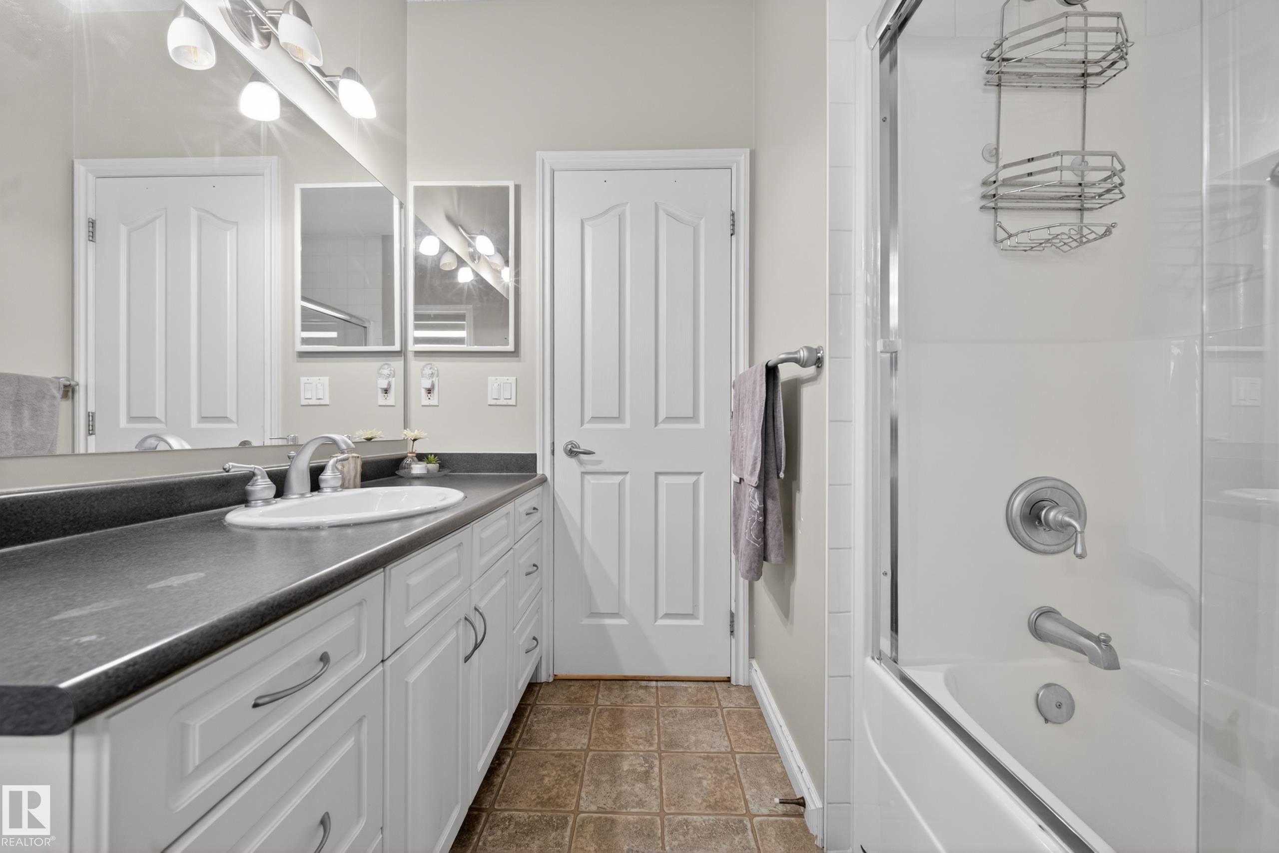Spacious bathroom featuring a single vanity with an integrated sink and dark countertop, white cabinetry with brushed nickel hardware, a full wall mirror, and a shower-over-tub combination with a glass enclosure - 4113 59 Street, Beaumont, AB