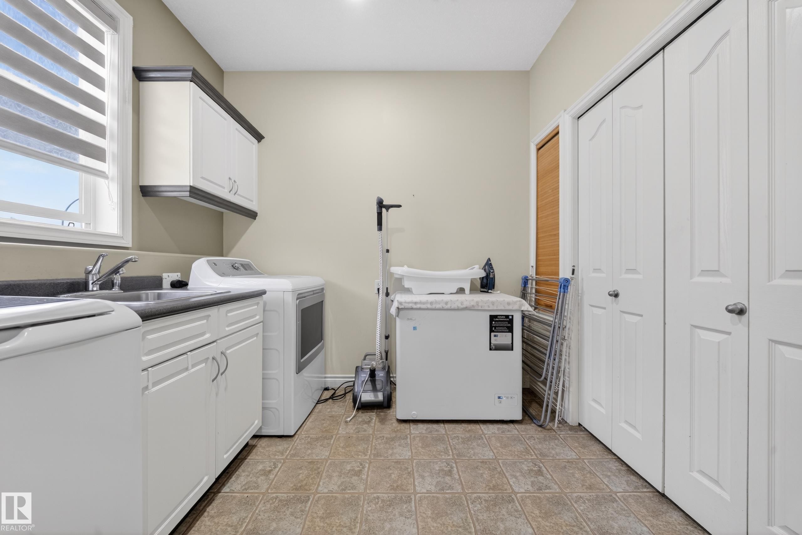 Dedicated laundry room featuring a utility sink, dark countertop, white cabinetry with silver hardware, and tiled flooring - 4113 59 Street, Beaumont, AB