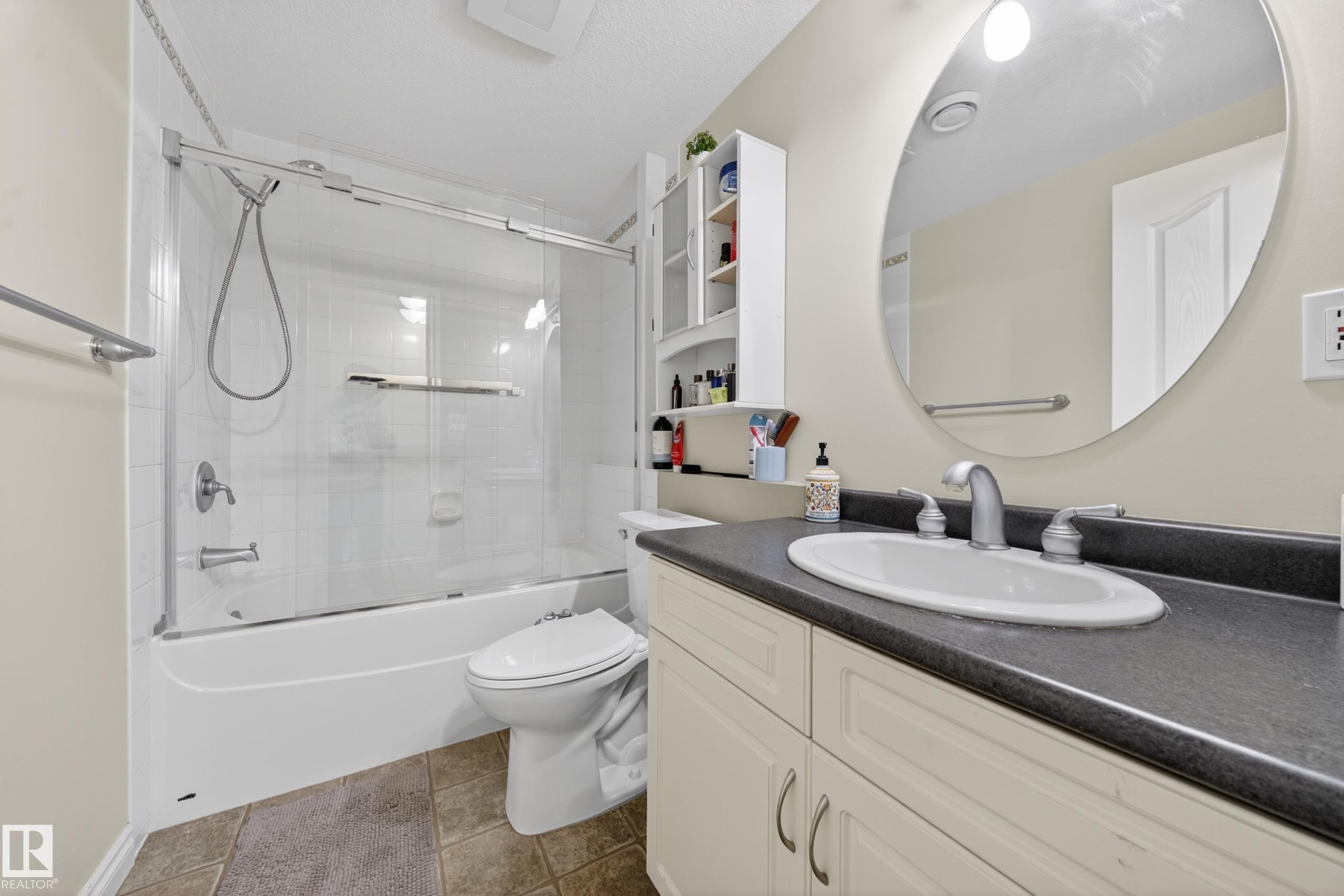 Bathroom featuring a combination tub and shower with glass enclosure, single vanity with integrated sink, dark countertop, and a large round wall mirror - 4113 59 Street, Beaumont, AB