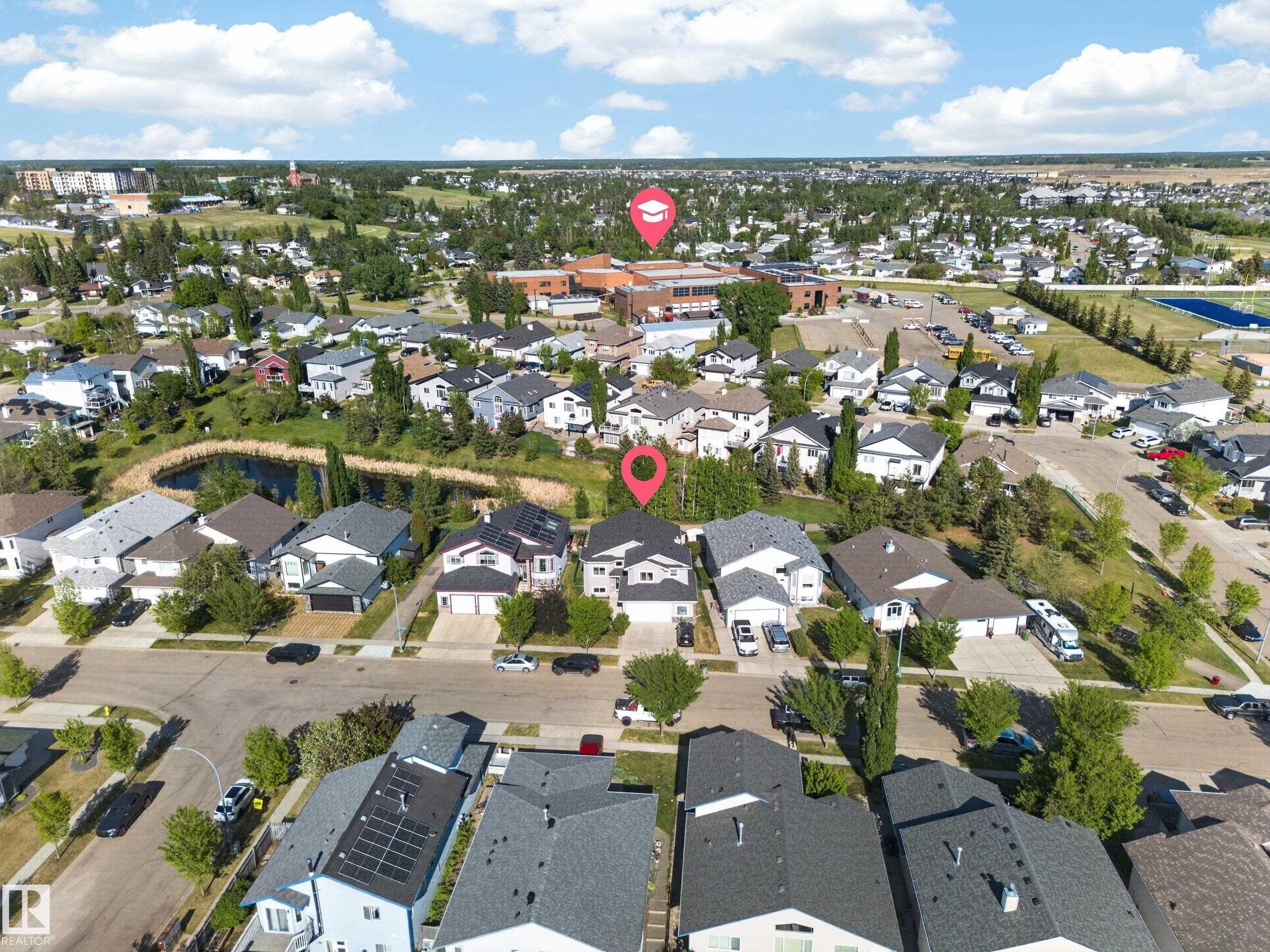 Aerial perspective showcasing a residential area with a neighborhood pond, mature trees, and nearby community buildings - 4113 59 Street, Beaumont, AB