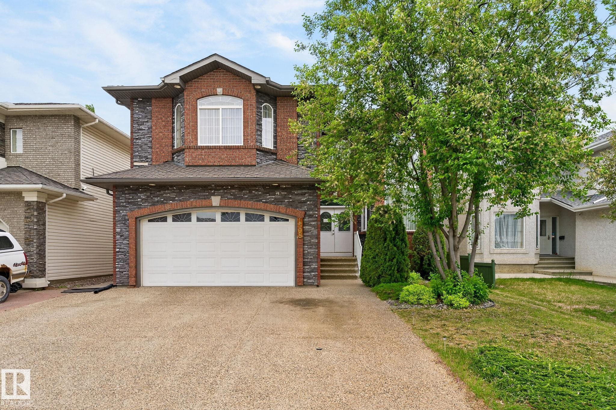 Brick and stone facade residence featuring an arched garage door, decorative window accents, a covered entry, an exposed aggregate driveway, and mature landscaping - 1508 Palmer Close, Edmonton, AB