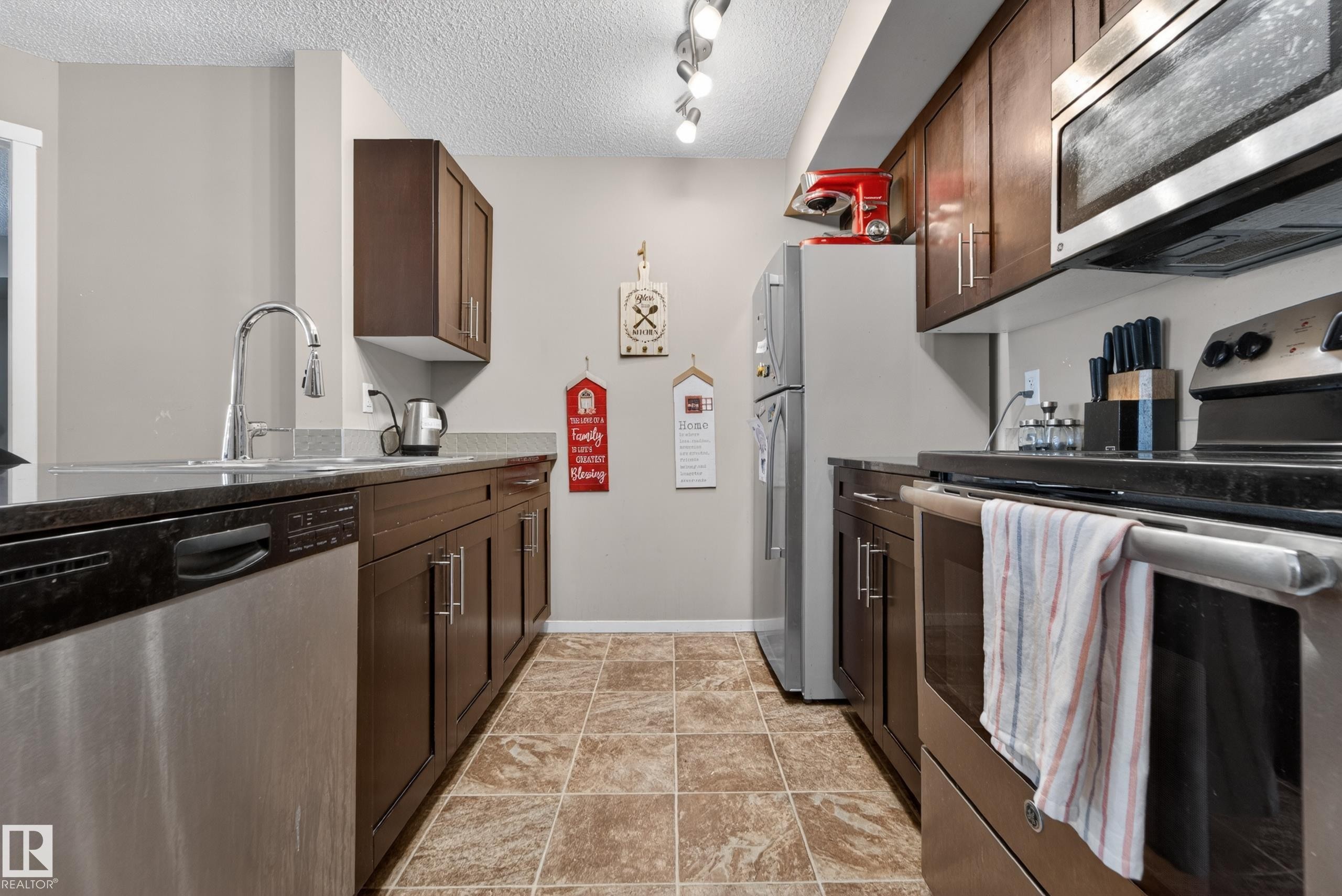 Galley kitchen featuring dark wood cabinetry, stainless steel appliances, and a tiled floor - 225 1510 Watt Drive, Edmonton, AB - Indoor Photo Showing Kitchen