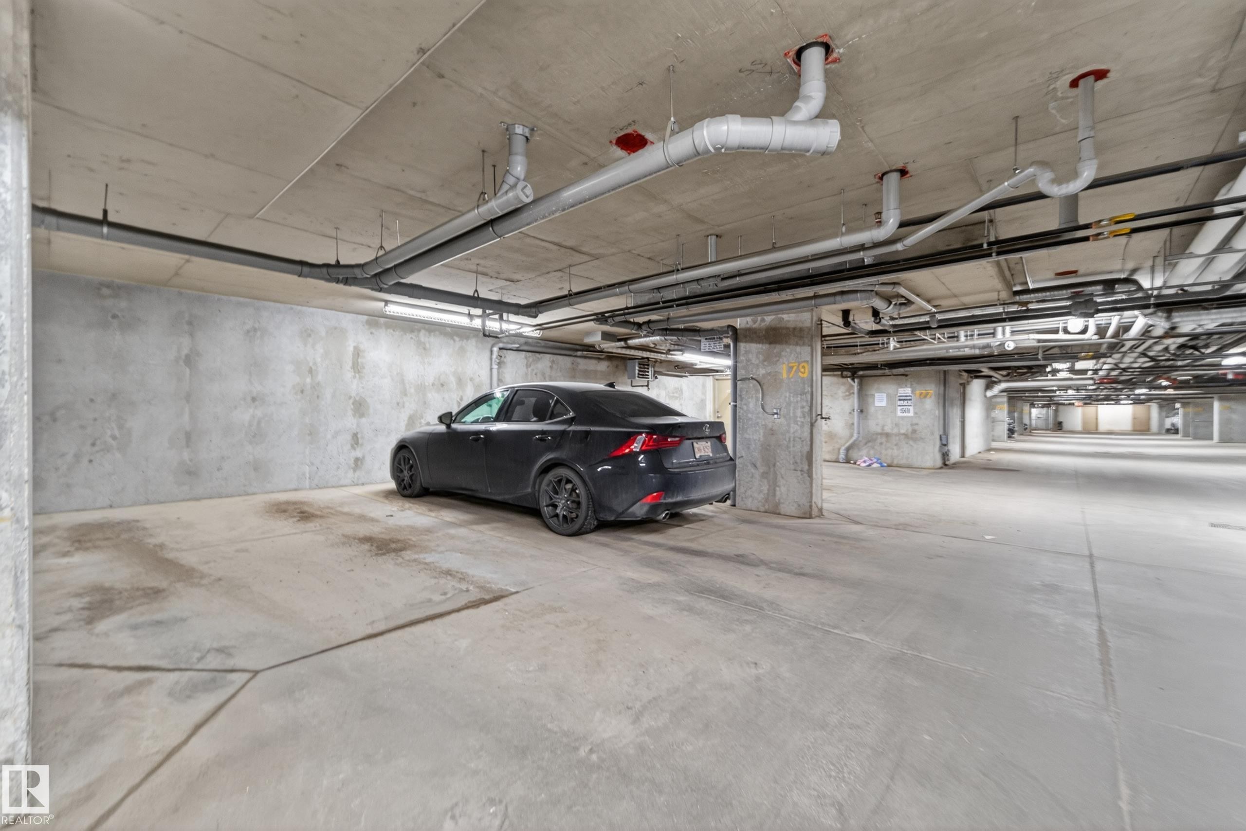 Spacious concrete parking garage featuring exposed overhead piping, concrete columns, and ample lighting - 225 1510 Watt Drive, Edmonton, AB - Indoor Photo Showing Garage