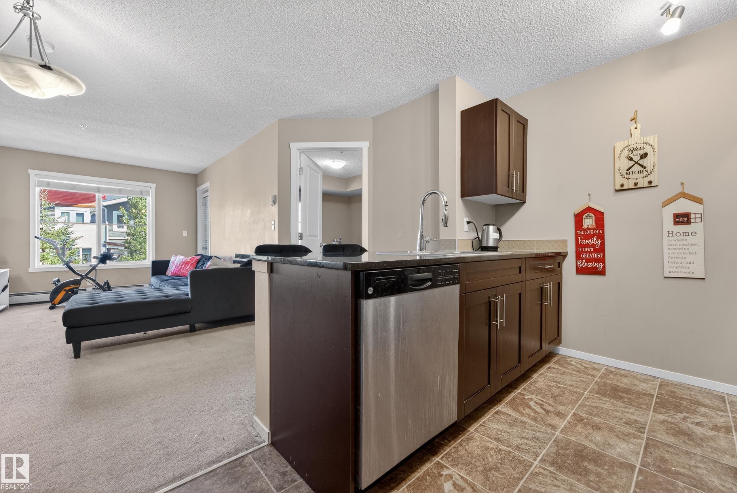 Contemporary kitchen area featuring wood-finish cabinetry, stainless steel dishwasher, and a dark stone countertop - 225 1510 Watt Drive, Edmonton, AB - Indoor