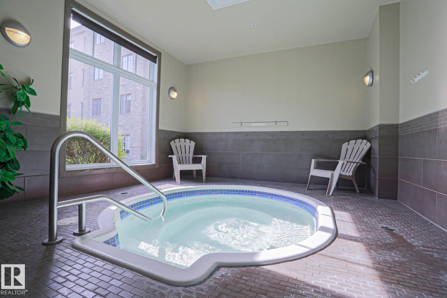 Indoor hot tub with a stainless steel grab rail, mosaic tile detailing, and a large window - 117 160 Magrath Road, Edmonton, AB - Indoor Photo Showing Other Room
