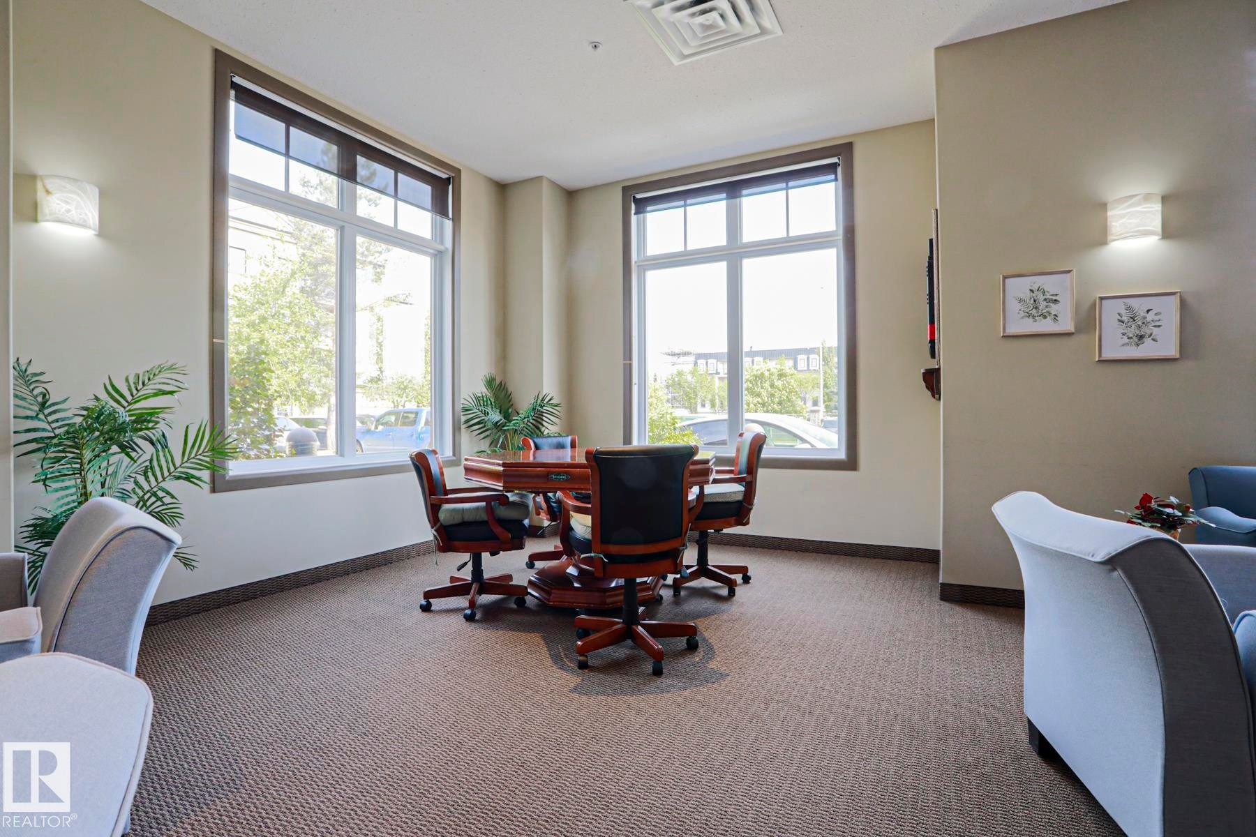 Spacious room featuring broad windows with roller shades, neutral tone walls, patterned carpet flooring, and integrated wall sconces - 117 160 Magrath Road, Edmonton, AB - Indoor Photo Showing Office
