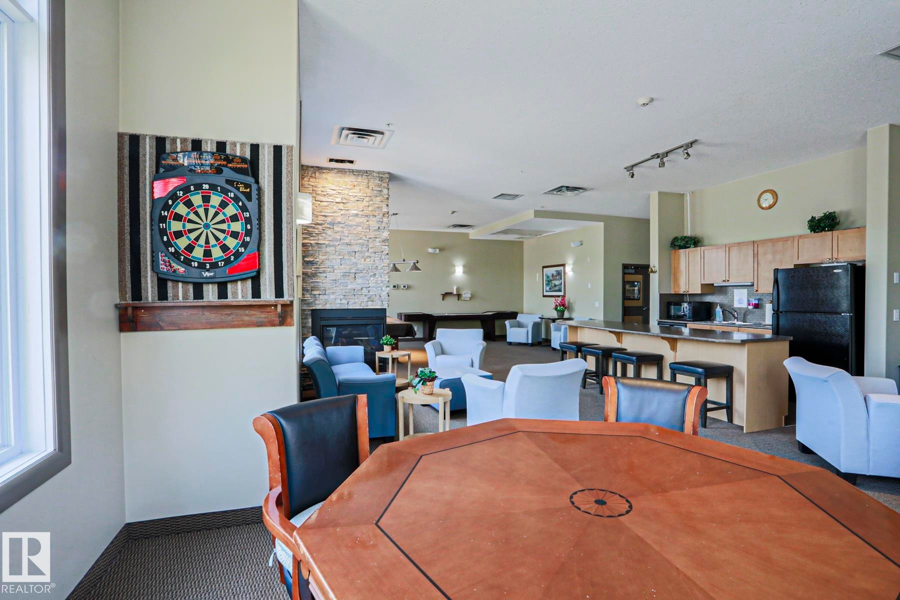 Spacious communal area featuring a built-in kitchen with wood cabinetry and a breakfast bar, a stone accent wall with a fireplace, track lighting, and carpeted flooring - 117 160 Magrath Road, Edmonton, AB - Indoor Photo Showing Dining Room
