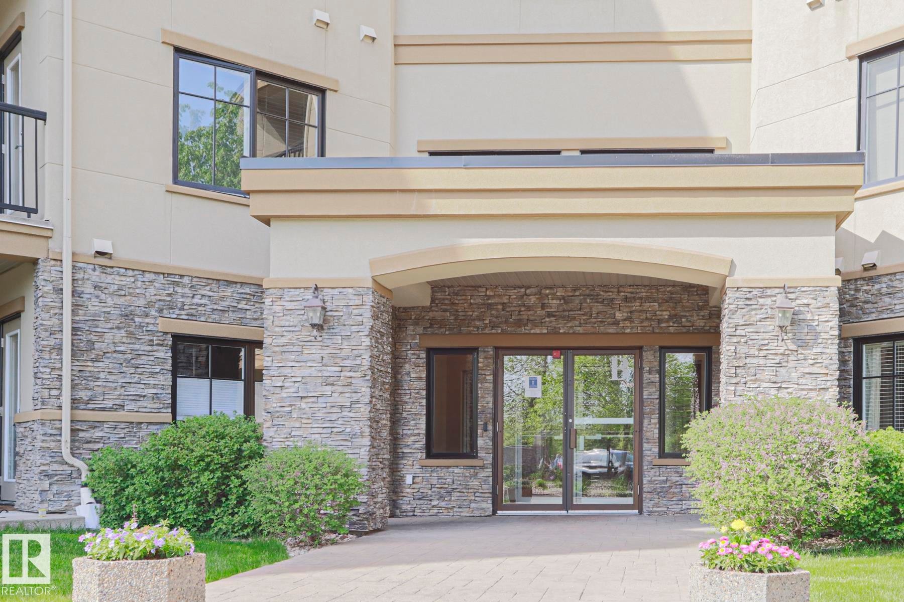 Building entrance featuring stacked stone facade, automatic glass doors, arched portico, perimeter landscaping, and paver walkway - 117 160 Magrath Road, Edmonton, AB - Outdoor With Exterior