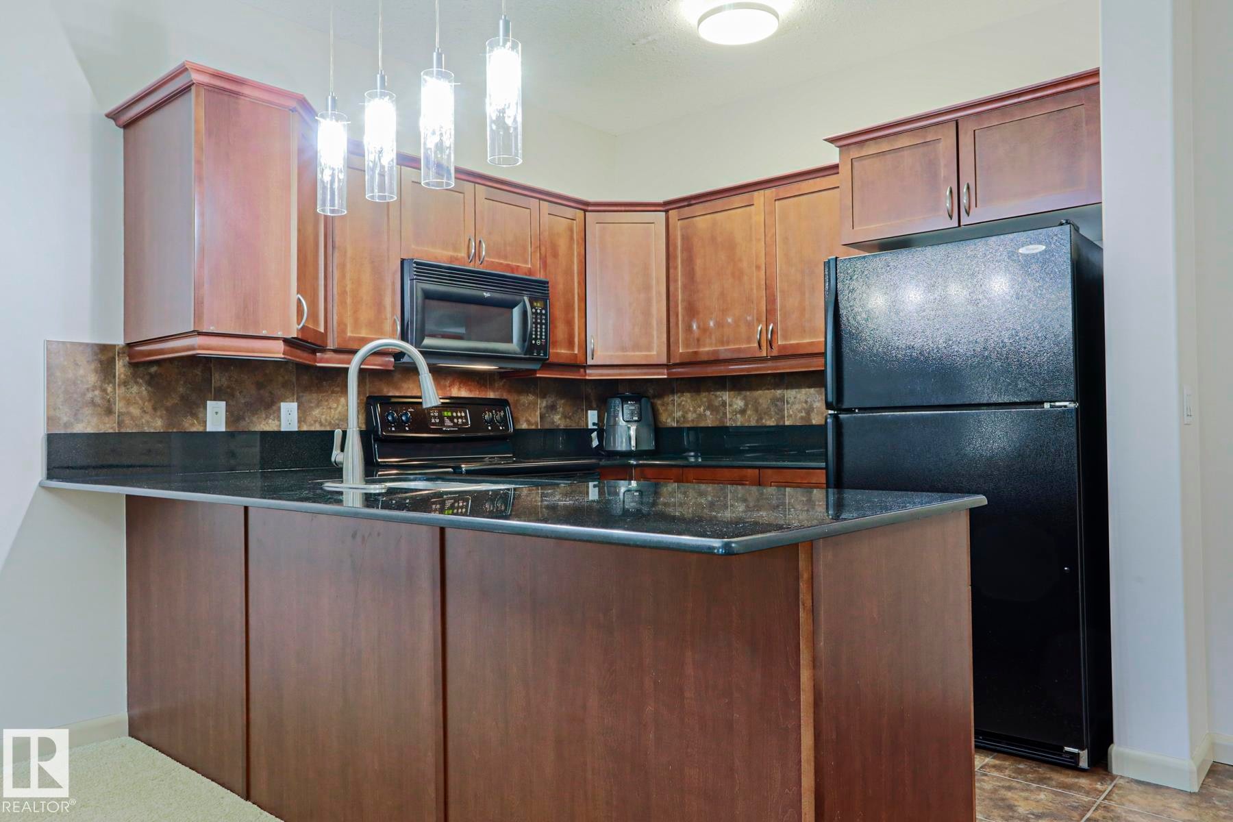 Kitchen with wood-finish cabinetry, black appliances, dark countertops, and a tiled backsplash - 117 160 Magrath Road, Edmonton, AB - Indoor Photo Showing Kitchen
