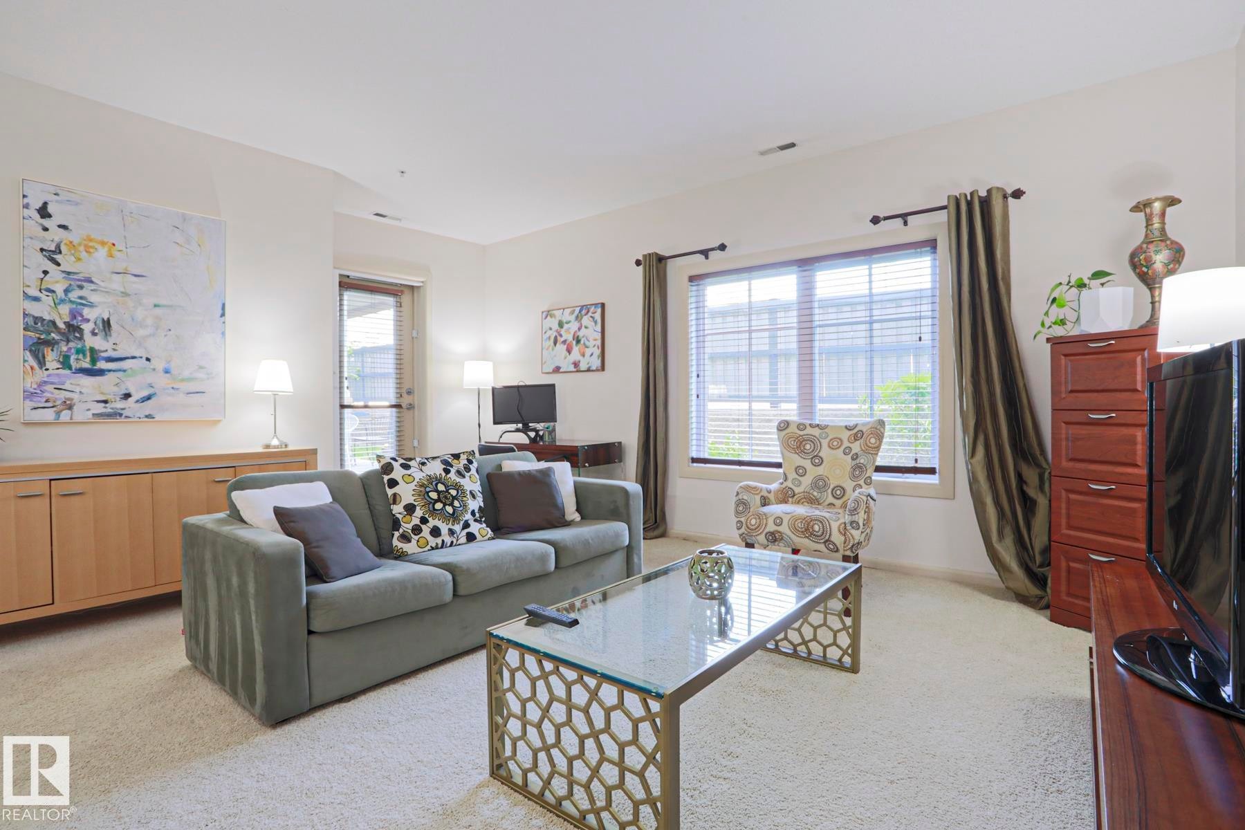 Living area featuring light-colored carpeting, a large window with blinds and drapes, and a glass-top coffee table with a geometric base - 117 160 Magrath Road, Edmonton, AB - Indoor Photo Showing Living Room