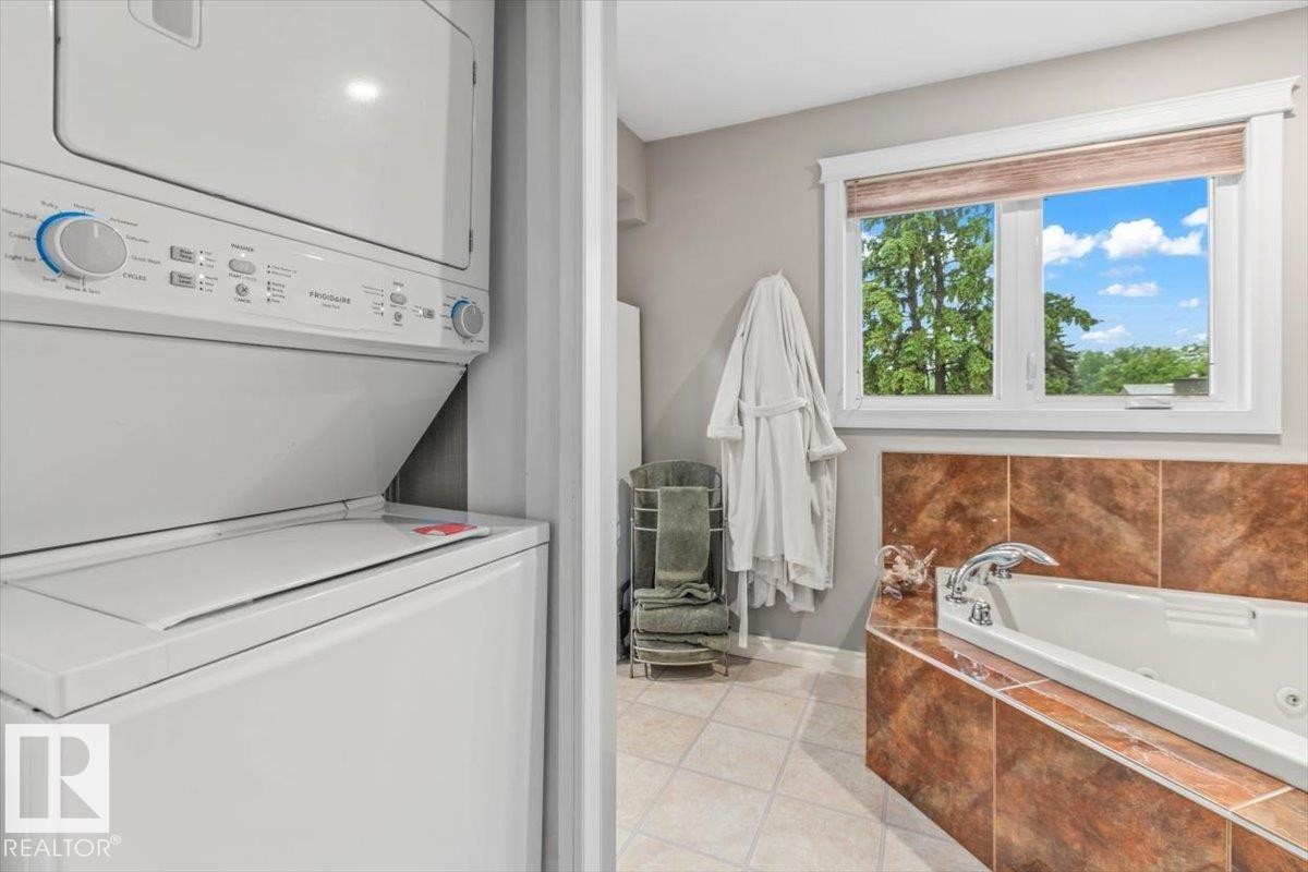 Integrated laundry appliances within a tiled bathroom featuring a built-in jetted tub, a window with exterior scenery, and light-toned floor tiles - 6720 29 Avenue, Edmonton, AB