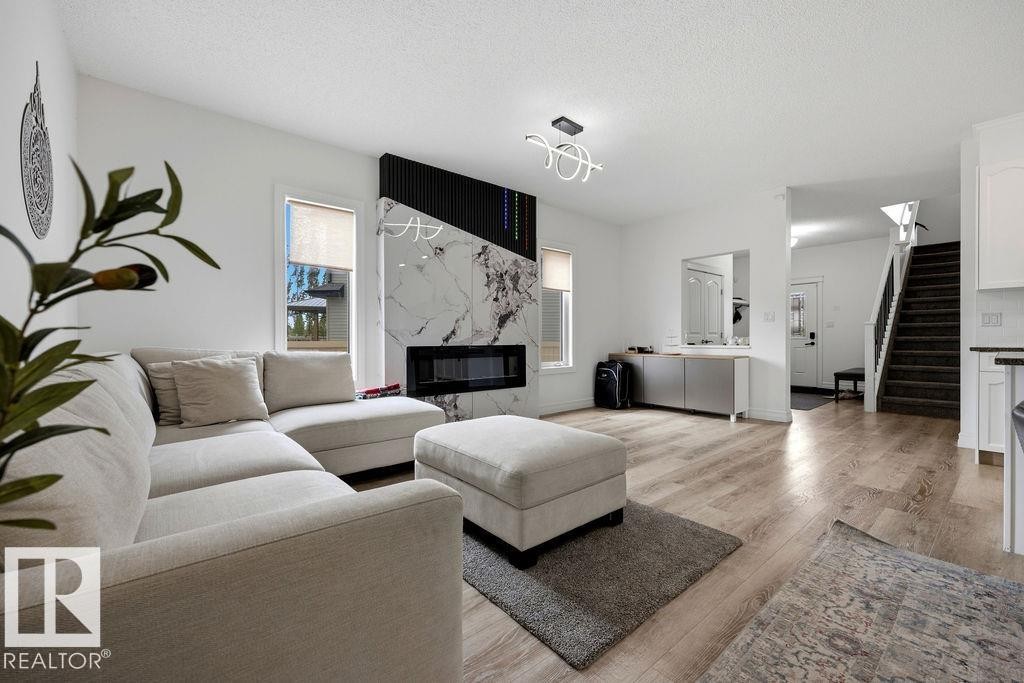 Spacious living area featuring a contemporary fireplace with a marble-finish surround and linear firebox, wood-finish flooring, a modern overhead light fixture, and a built-in storage cabinet - 13703 166 Avenue Nw, Edmonton, AB - Indoor Photo Showing Living Room With Fireplace