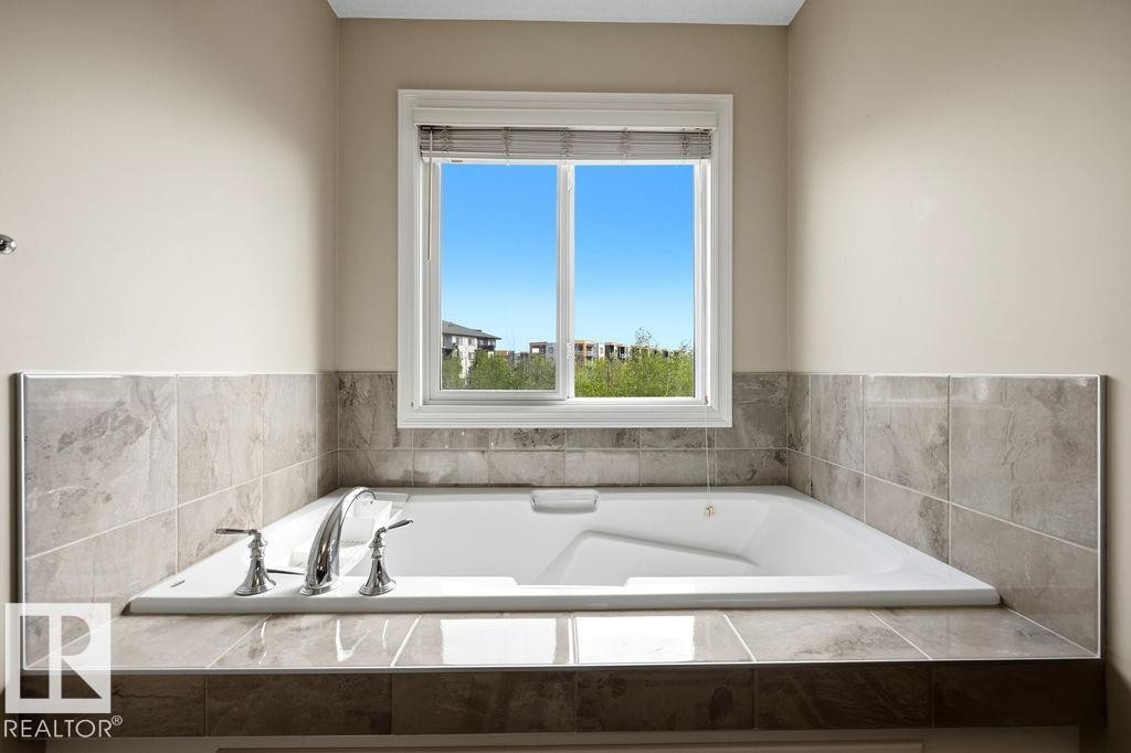 Built-in soaking tub with chrome fixtures, surrounded by large format tile in varying shades of gray, and a window with blinds - 13703 166 Avenue Nw, Edmonton, AB - Indoor Photo Showing Bathroom