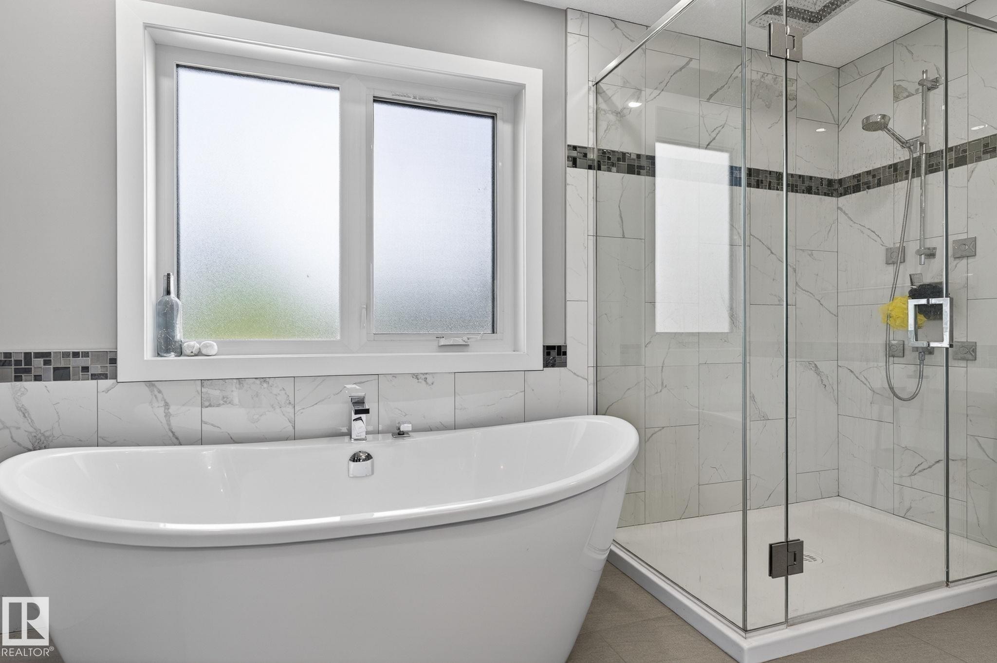 Spacious bathroom featuring a freestanding soaking tub with deck-mounted chrome fixtures, a glass-enclosed shower with multiple showerheads, white-veined tile walls, and a decorative mosaic accent band - 8809 148 Street, Edmonton, AB - Indoor Photo Showing Bathroom